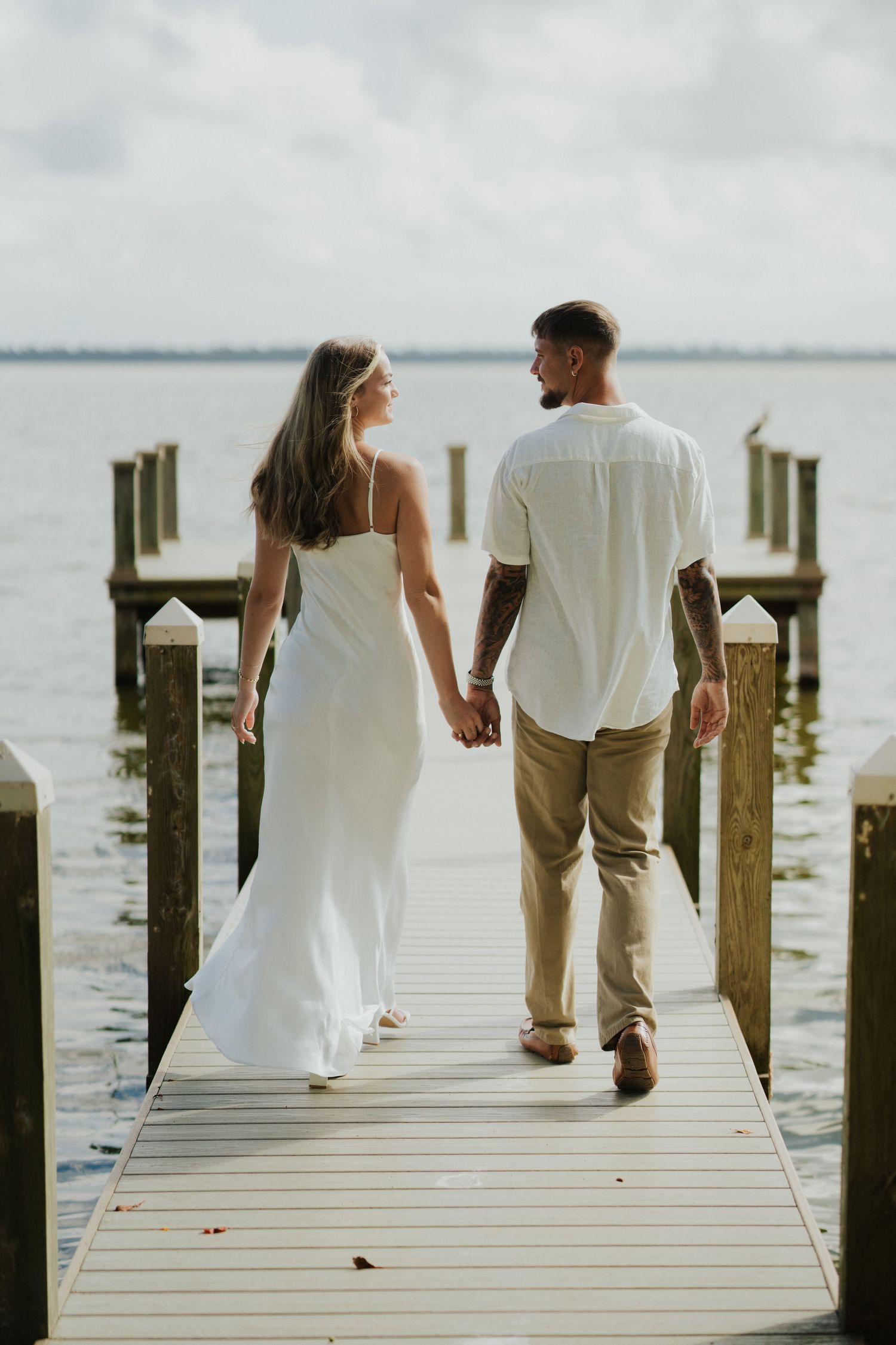 A couple in white attire walking together on a wooden dock by the waterfront at sunset.
