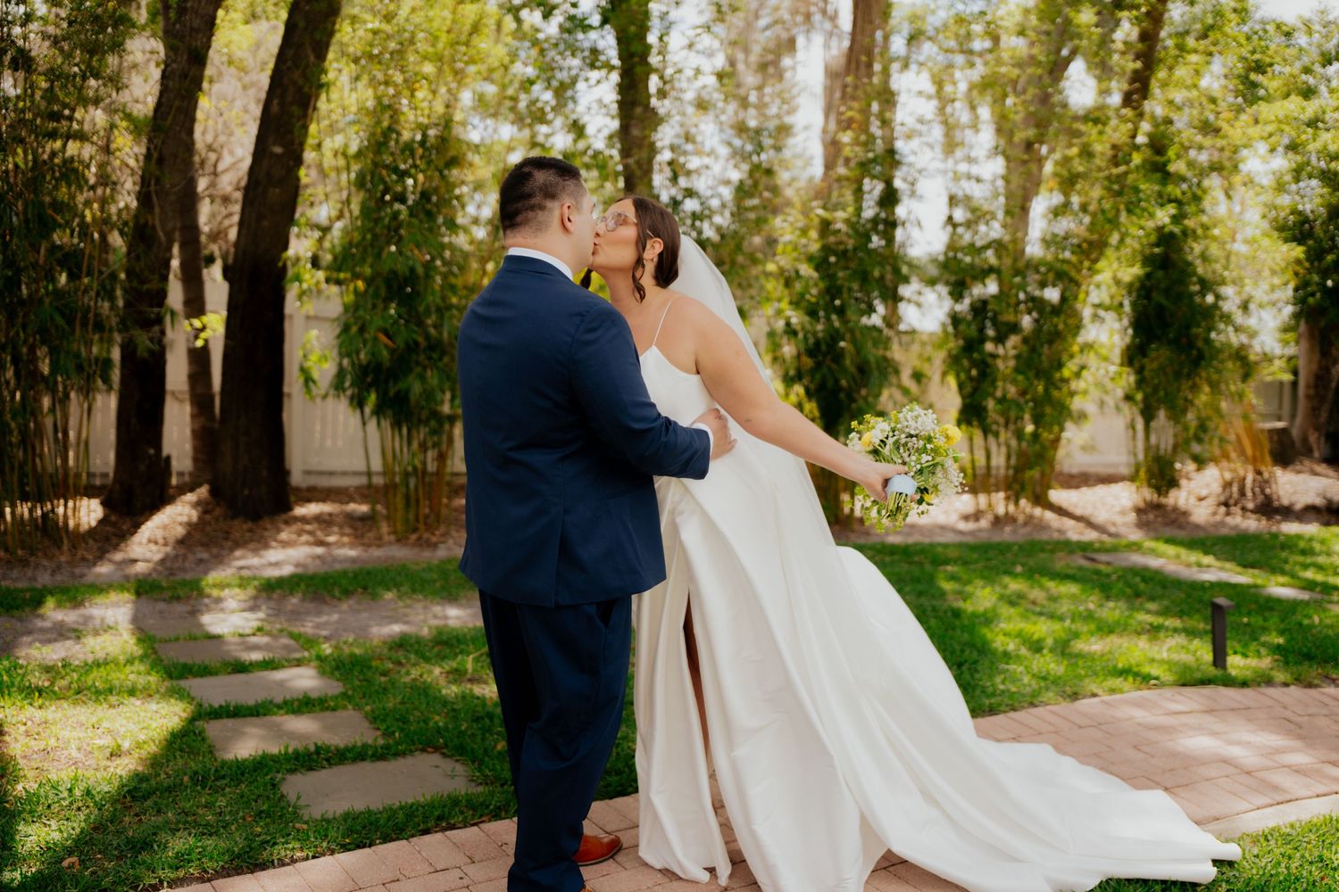 A couple in wedding attire sharing an intimate moment on a garden path surrounded by sunlit trees.