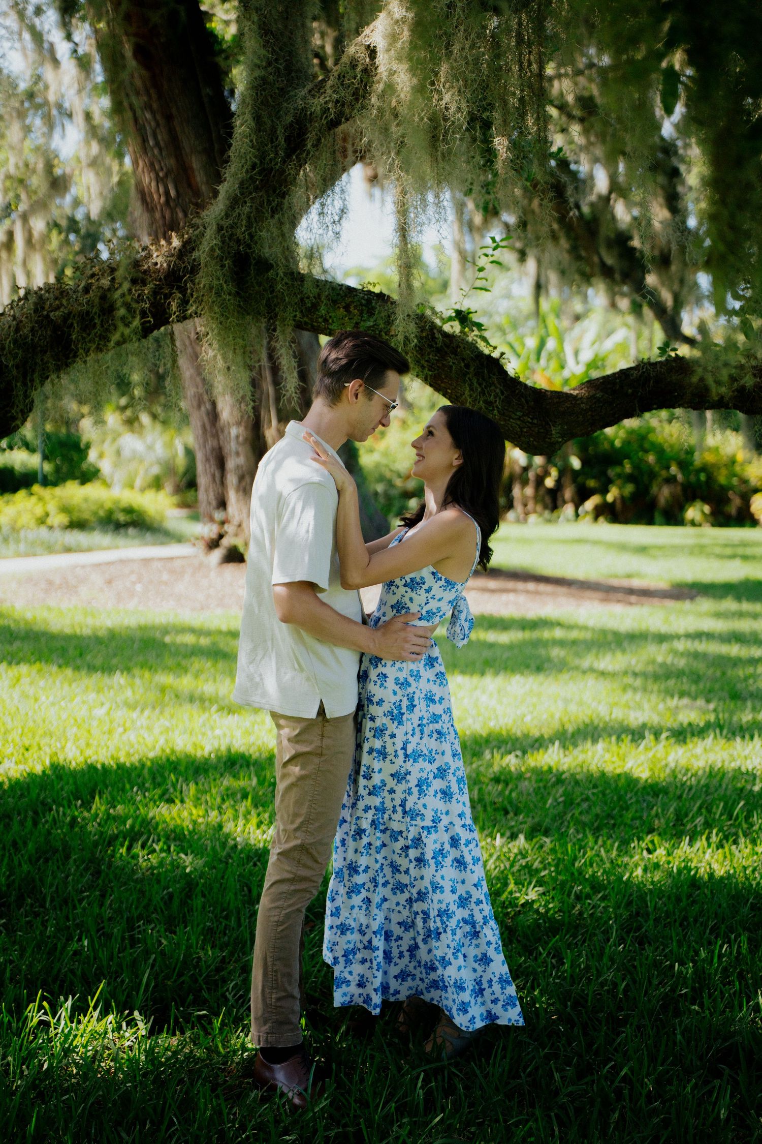 A romantic couple shares tender moments under moss-draped trees in a lush green park setting.