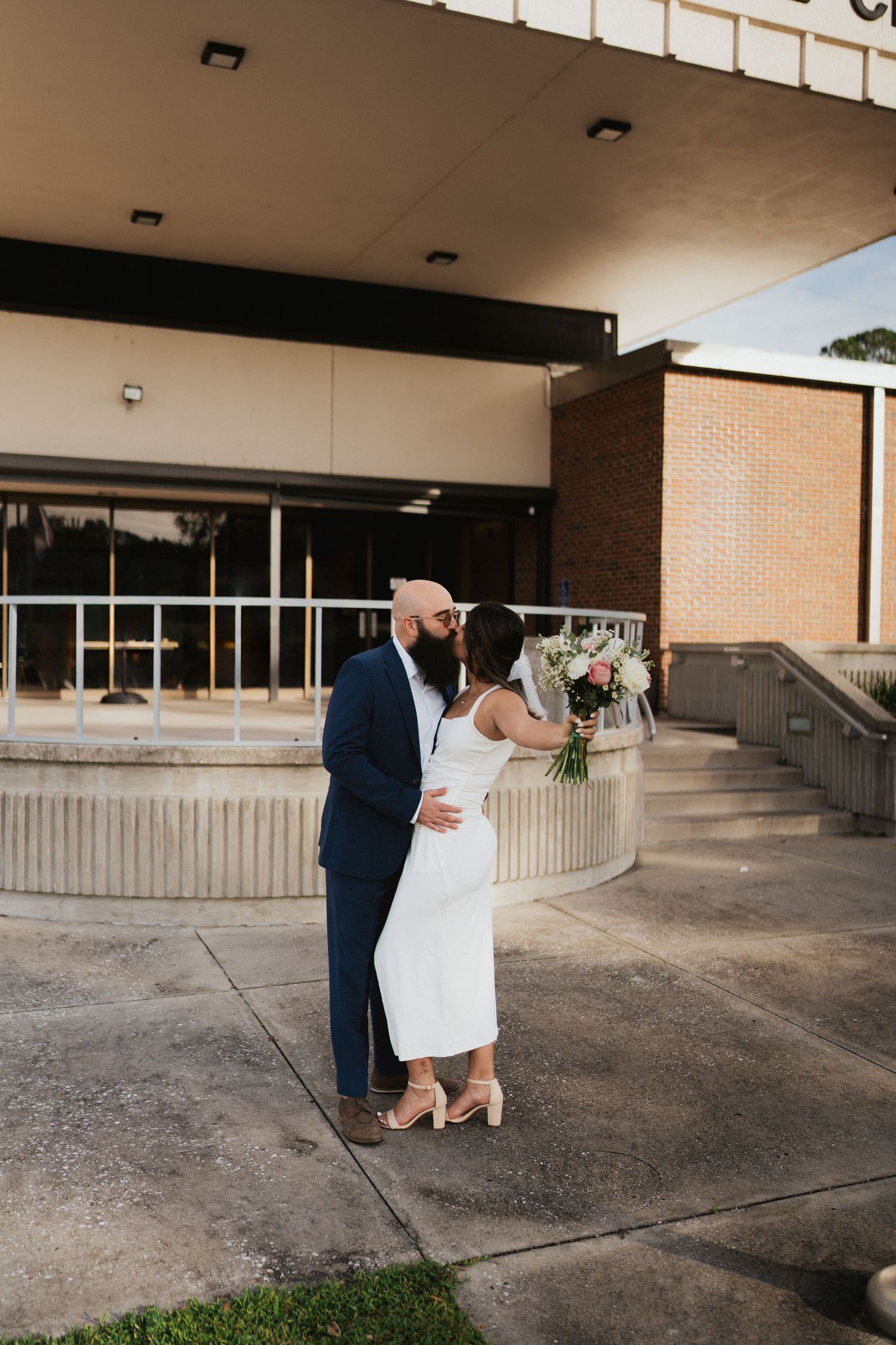 A couple in wedding attire standing outside a modern building entrance.