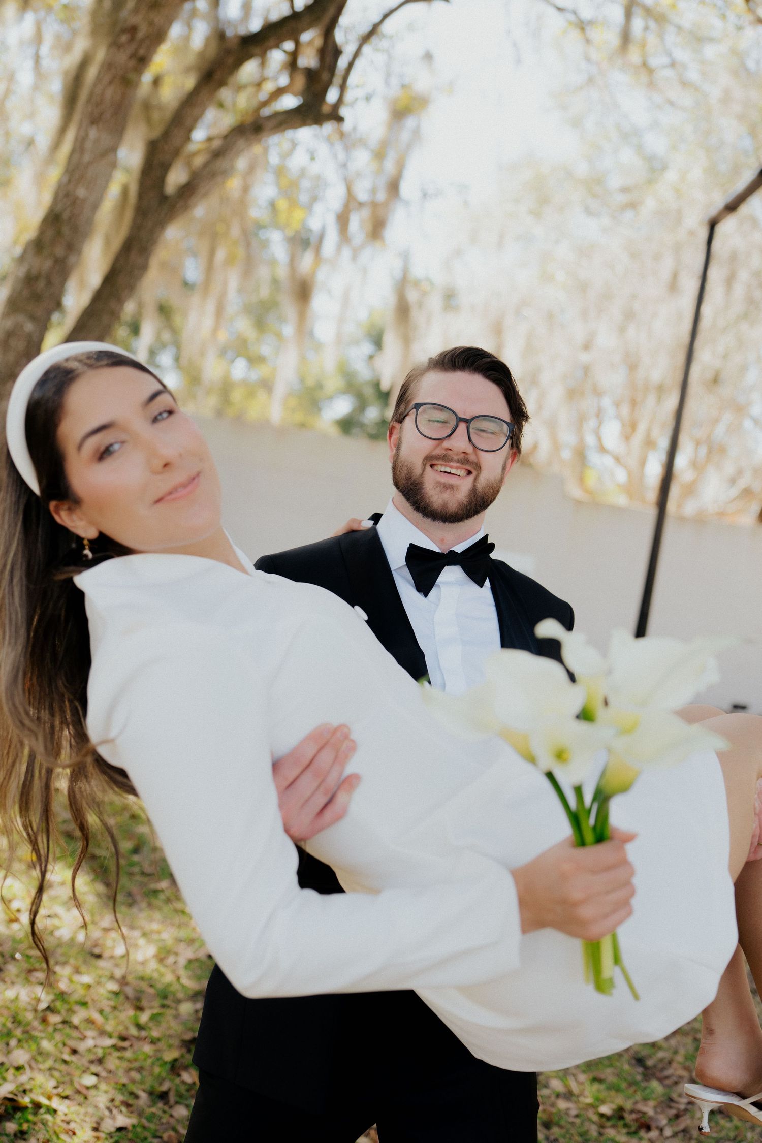 A couple in wedding attire share playful moments together outdoors among autumn trees.