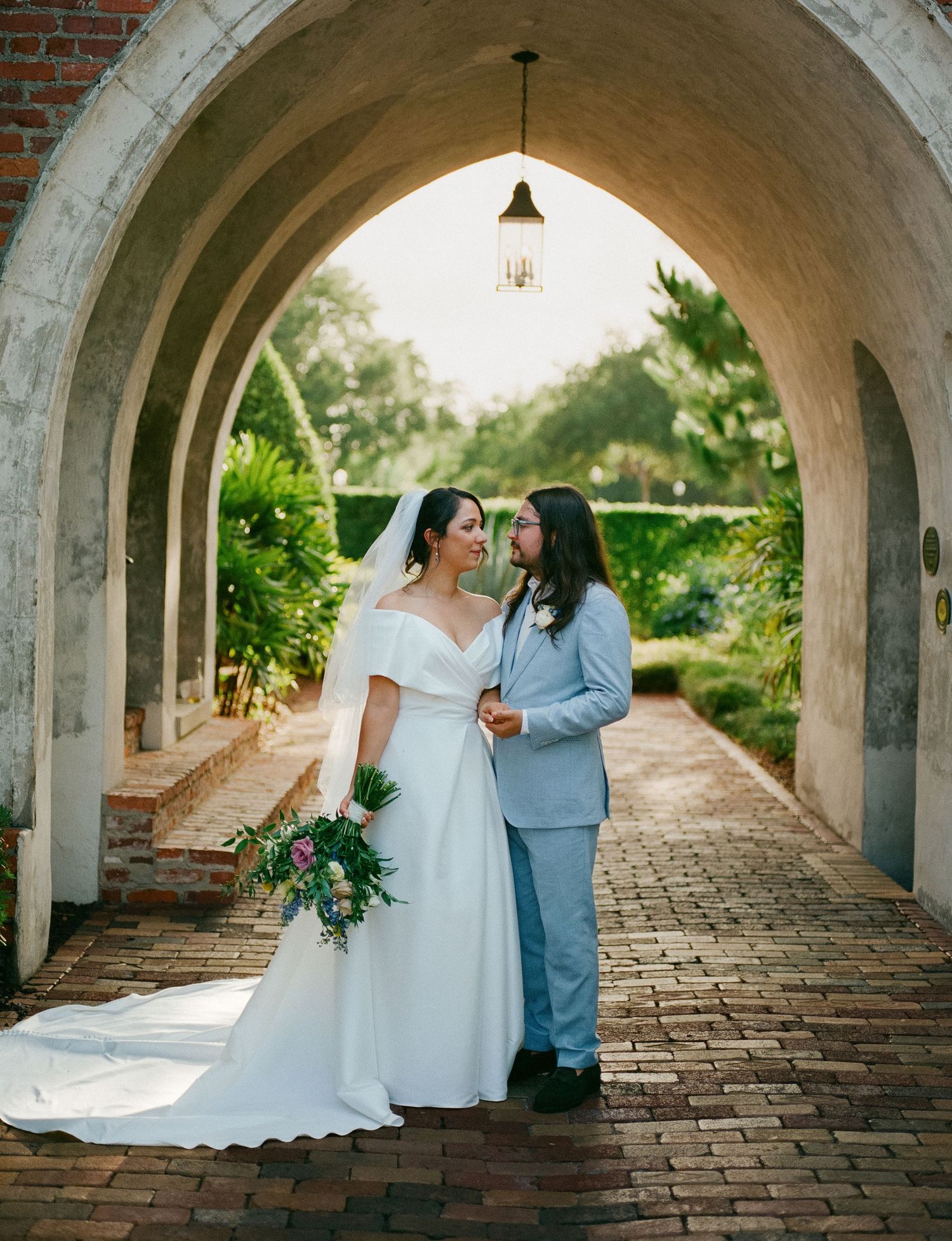 Bride and groom sharing a romantic moment under a stone archway on a brick pathway.