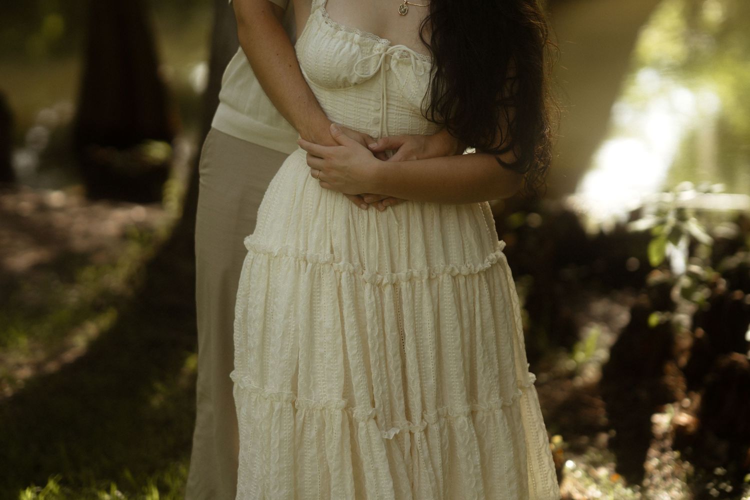Woman in flowing cream tiered dress standing in dappled sunlight in a lush garden setting.