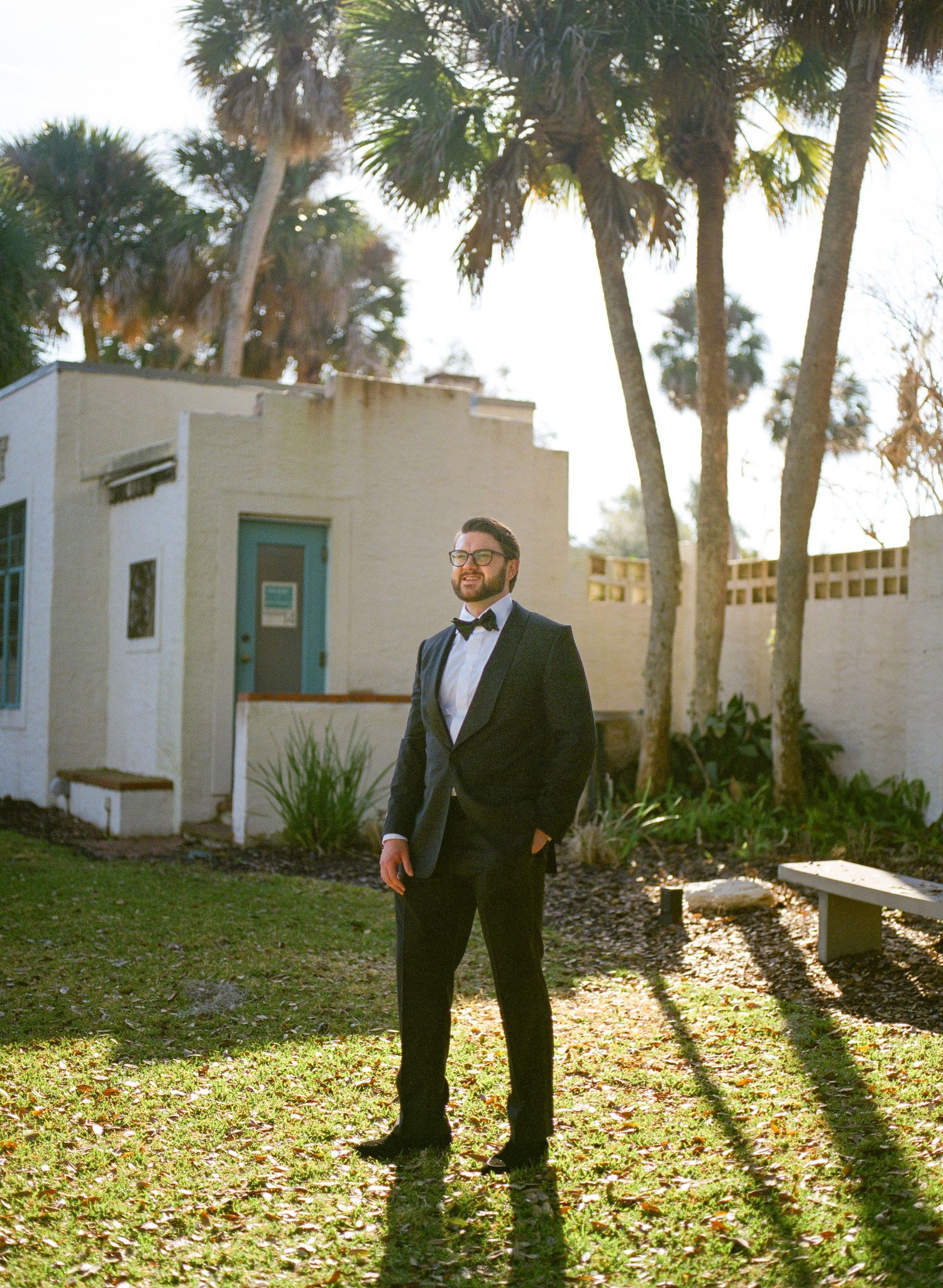 Dapper groom in black tuxedo with bow tie standing outdoors near palm trees in golden sunlight.