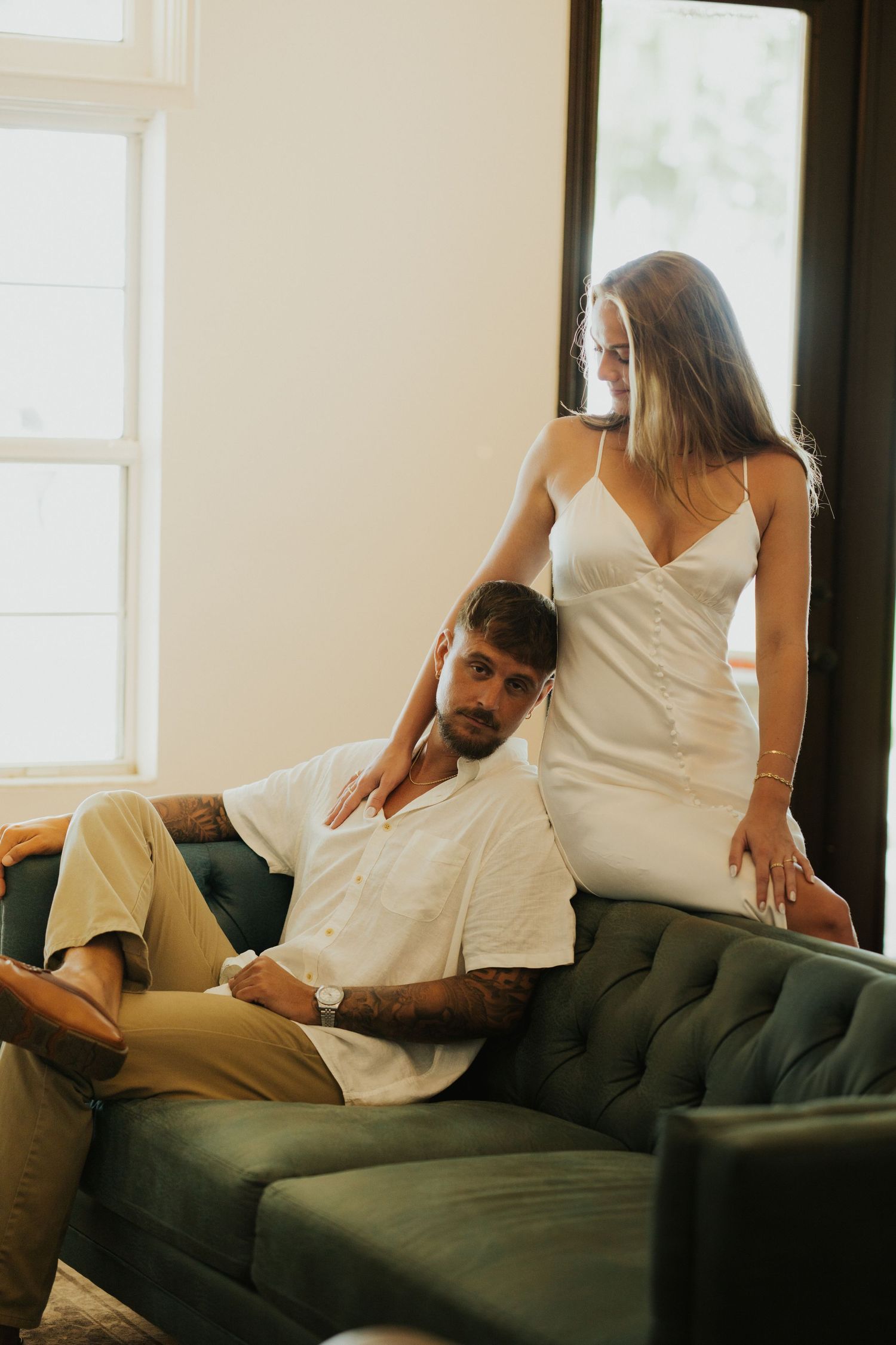 A couple in white clothing shares intimate moments on a dark gray couch in a bright, sunlit room.