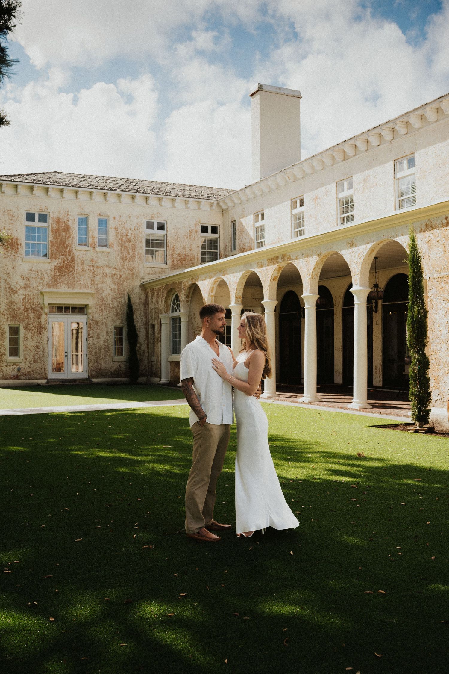 A couple dances on a manicured lawn surrounded by elegant stone archways and columns of a historic building.