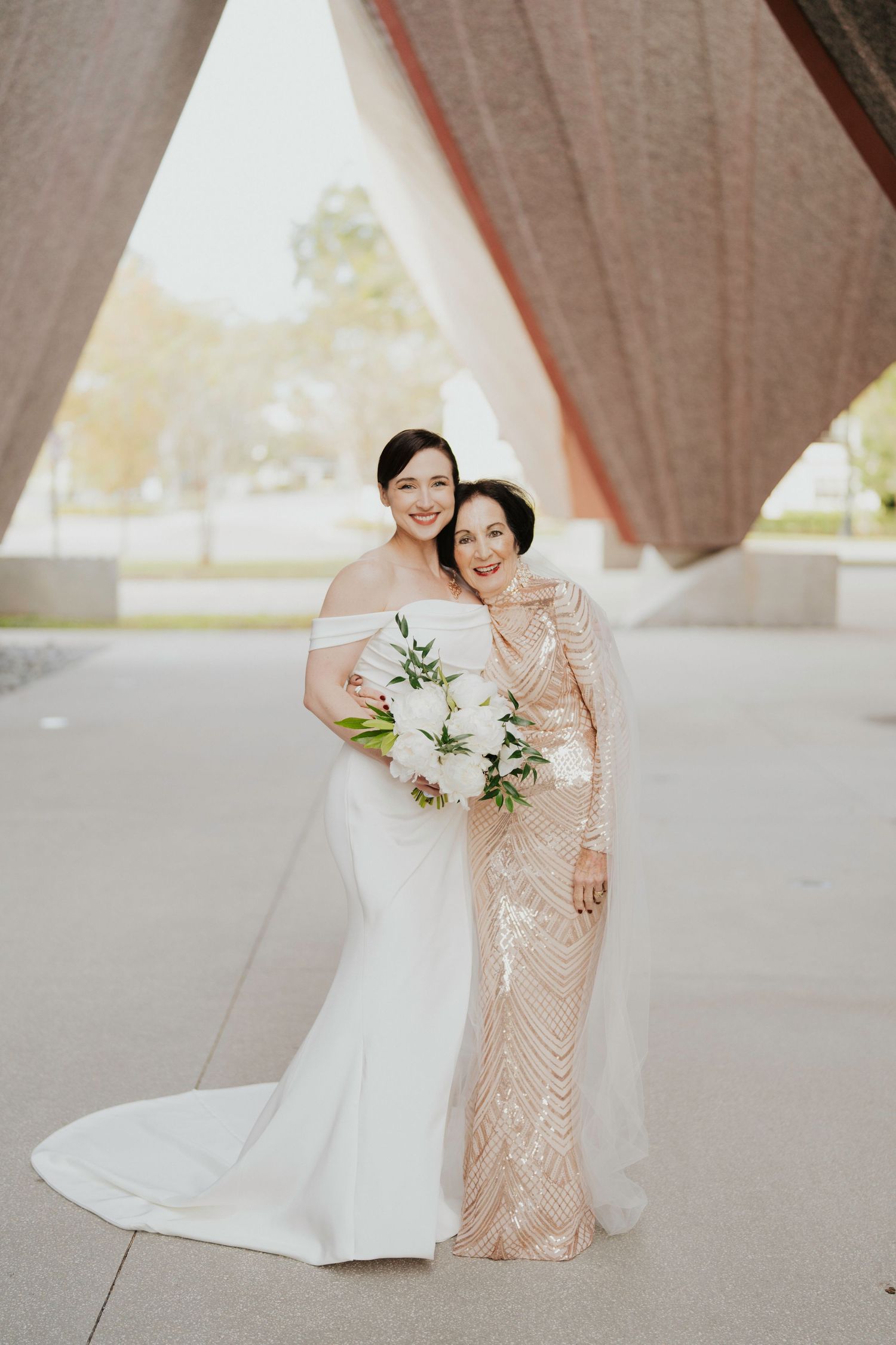 Wedding portrait showing bride in white gown and companion in blush sequined dress holding floral bouquets.