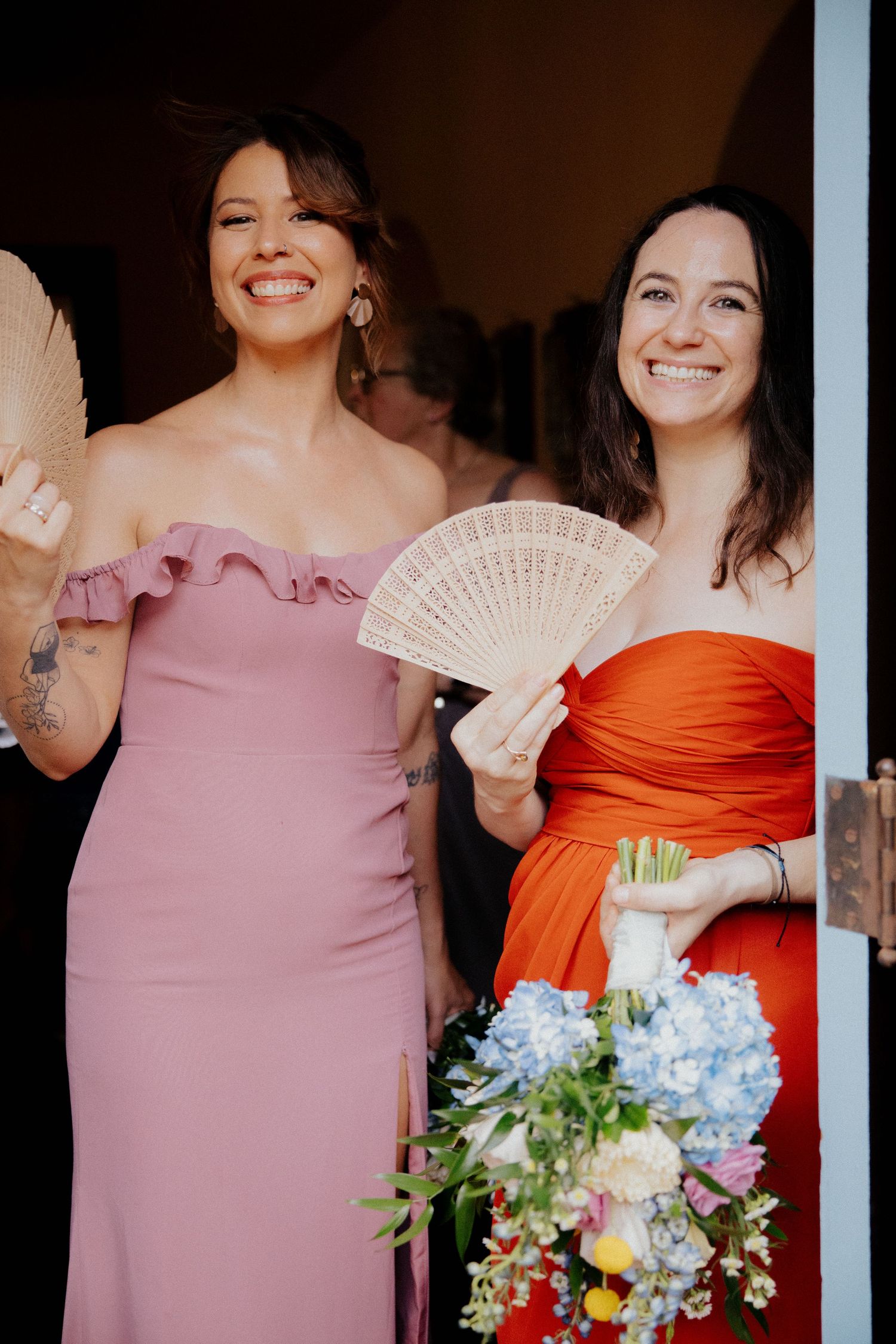 Wedding party members in pink and orange dresses holding bouquets and decorative fans.