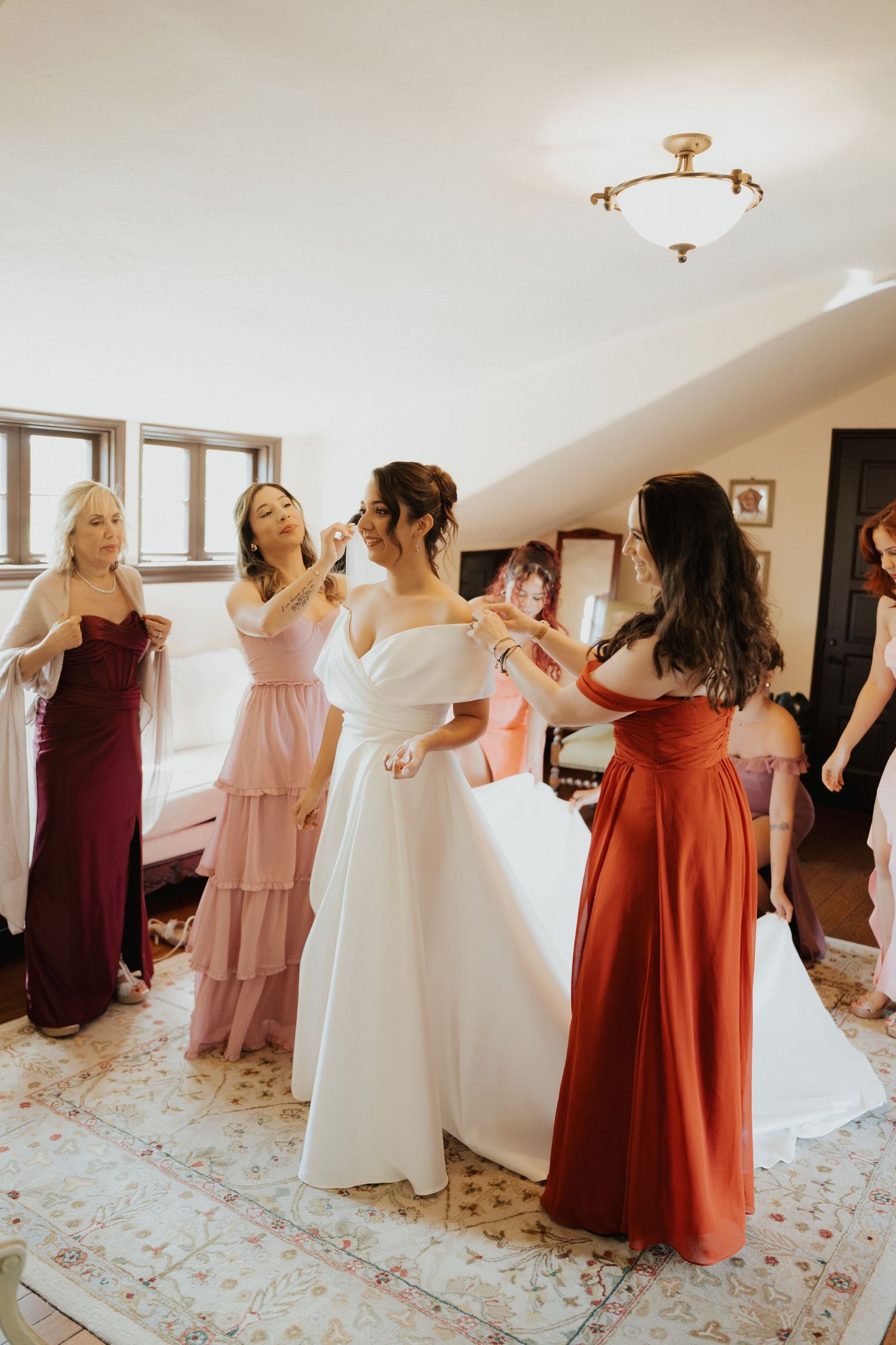 Bridesmaids in mixed pink and burgundy dresses dance and celebrate in a brightly lit room.
