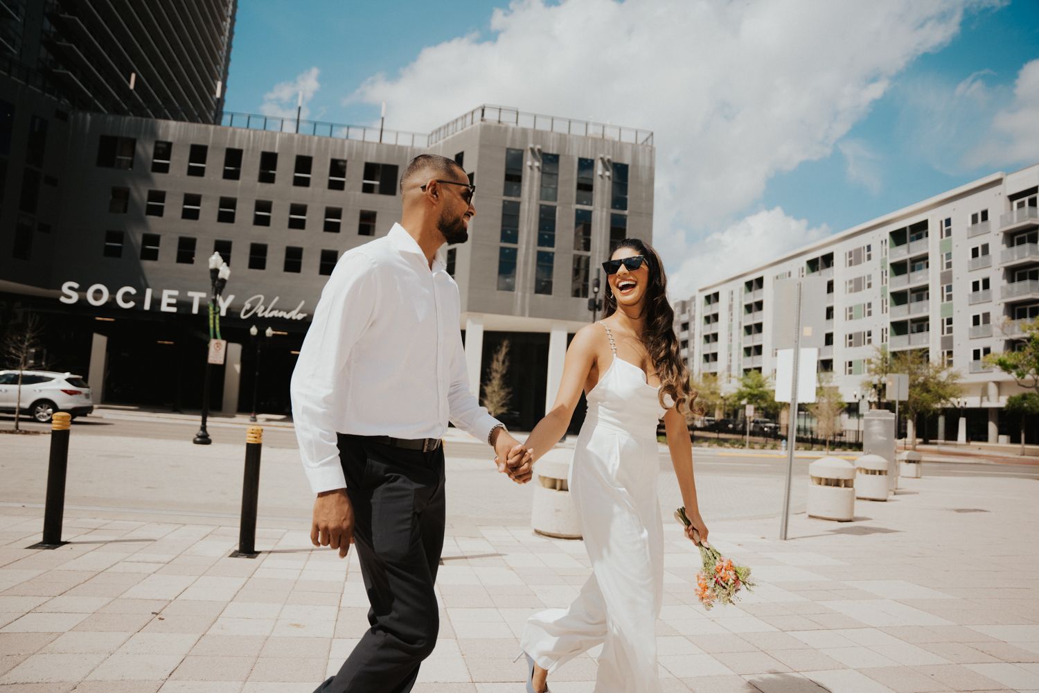 Newlyweds walk together in an urban setting with modern architecture and a 'Society' sign visible in background.