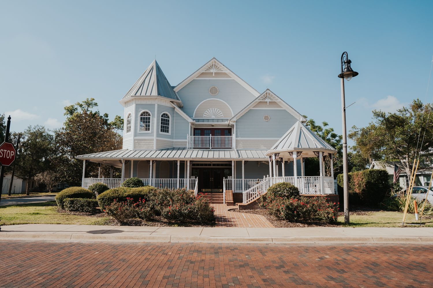 Elegant white Victorian-style building with wraparound porch and turret situated on a brick-paved street.