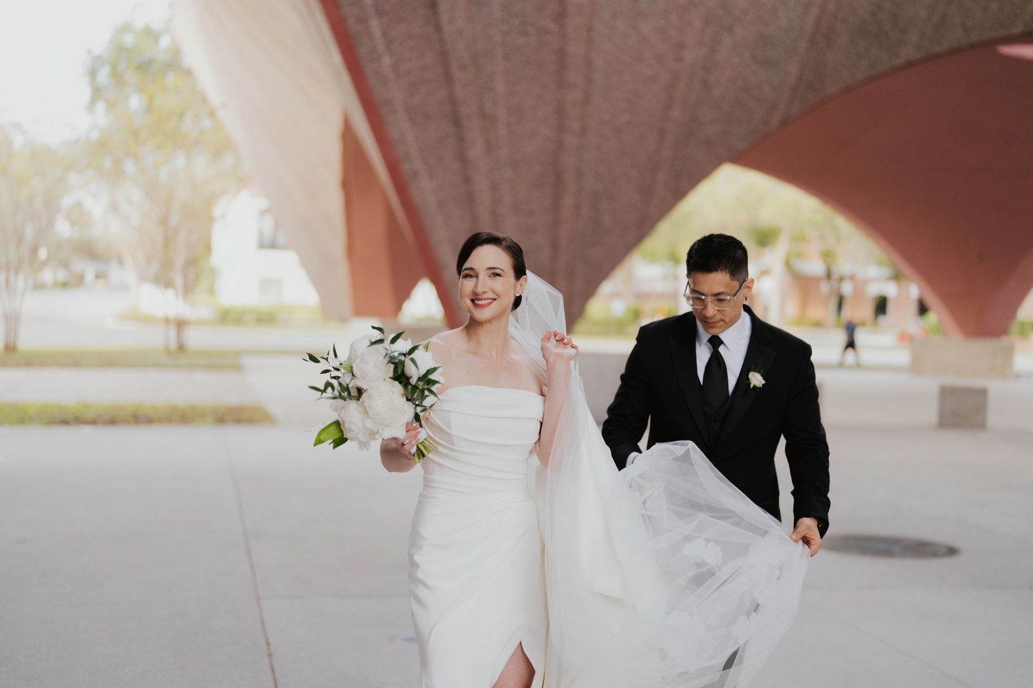 A couple in wedding attire runs joyfully under a curved architectural structure while holding a white bouquet.