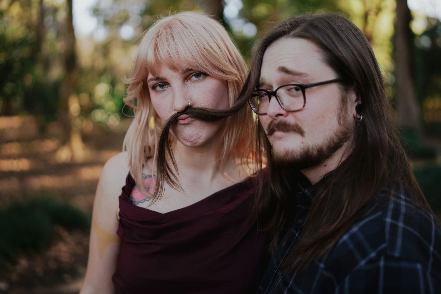 Two people making silly faces with hair as mustaches in a wooded setting.