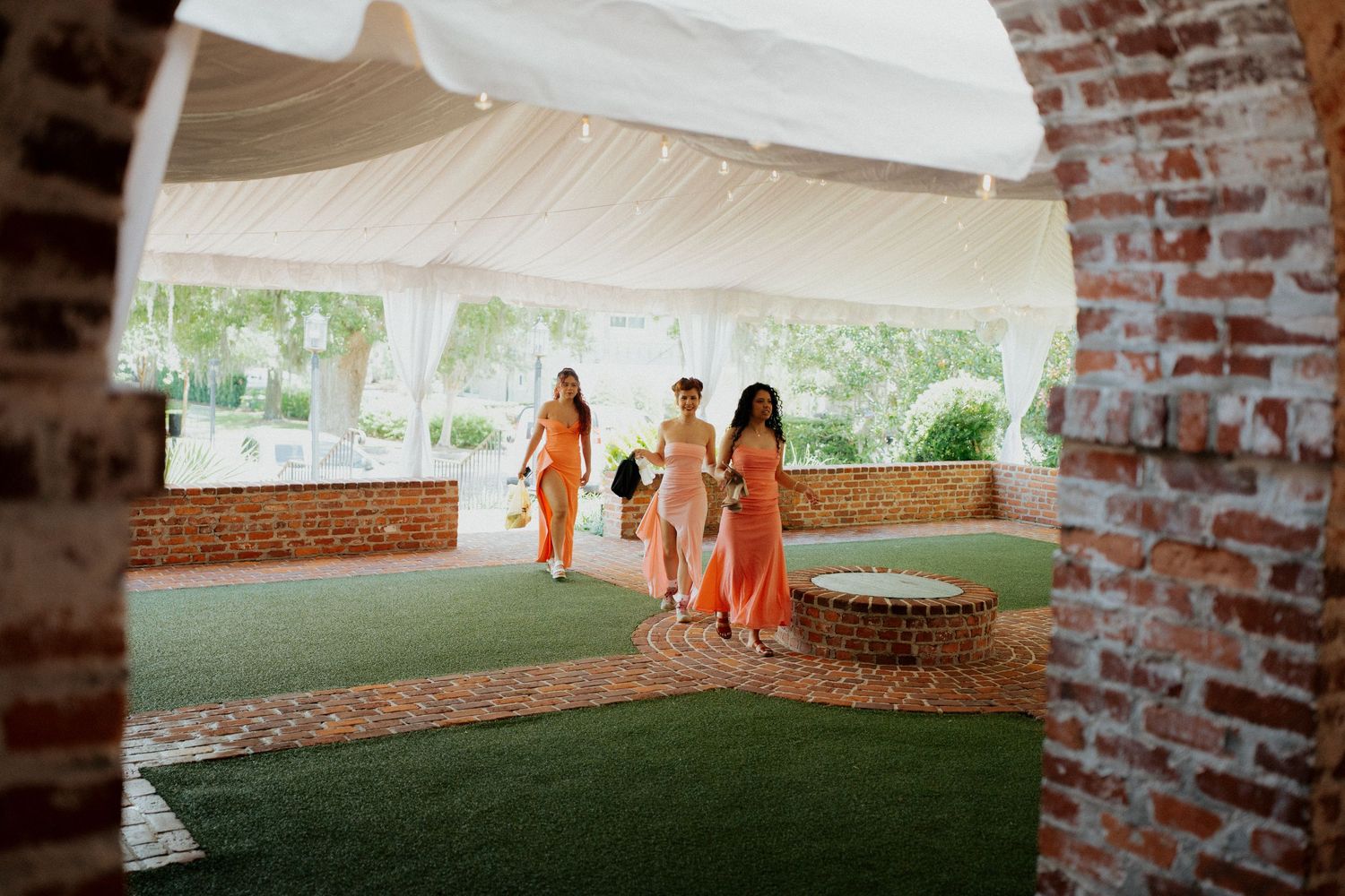 Orange-clad bridesmaids pose under a white tent canopy at an outdoor wedding venue.