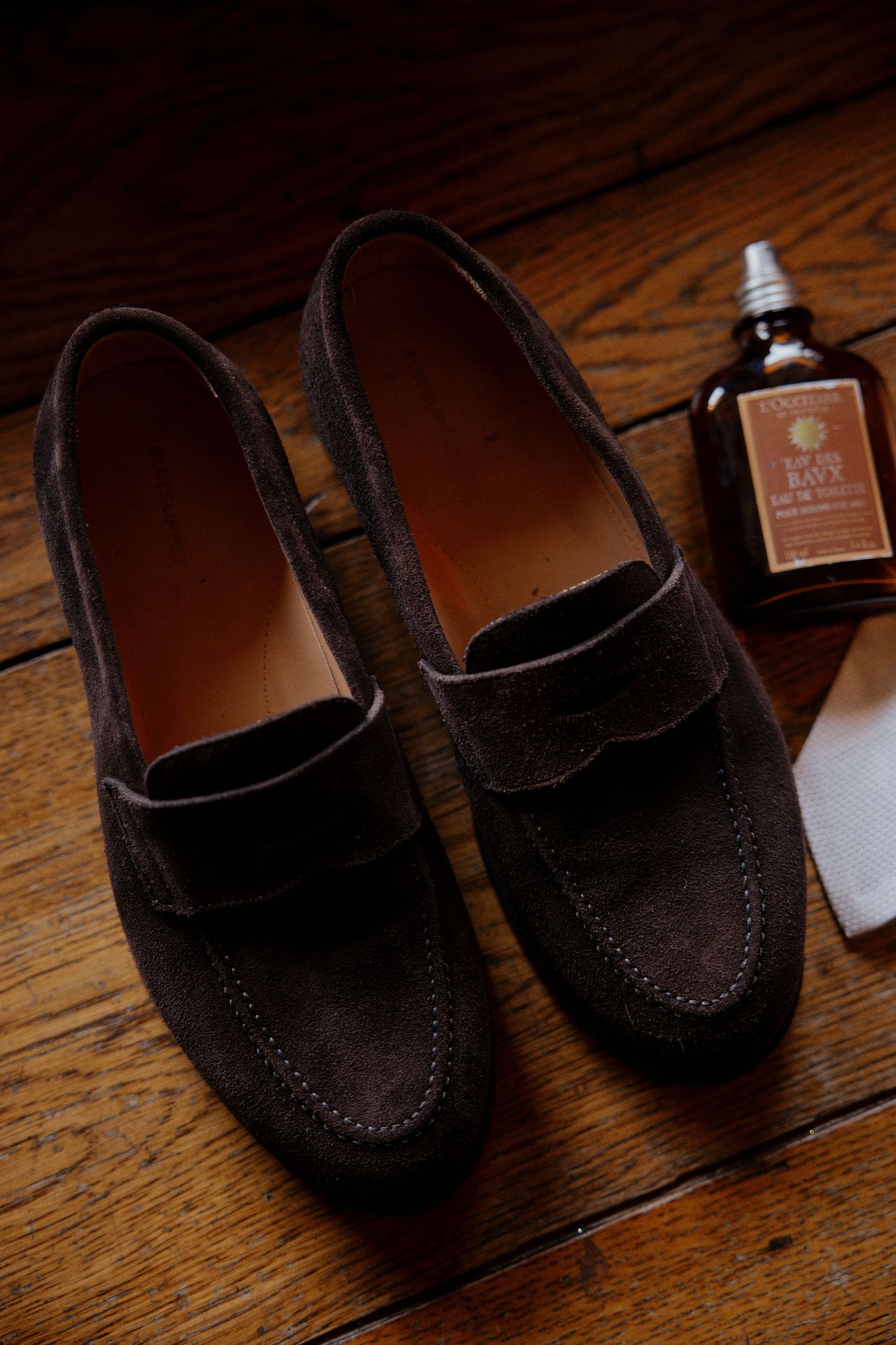 Dark brown suede loafers sit on a wooden surface in dramatic lighting.