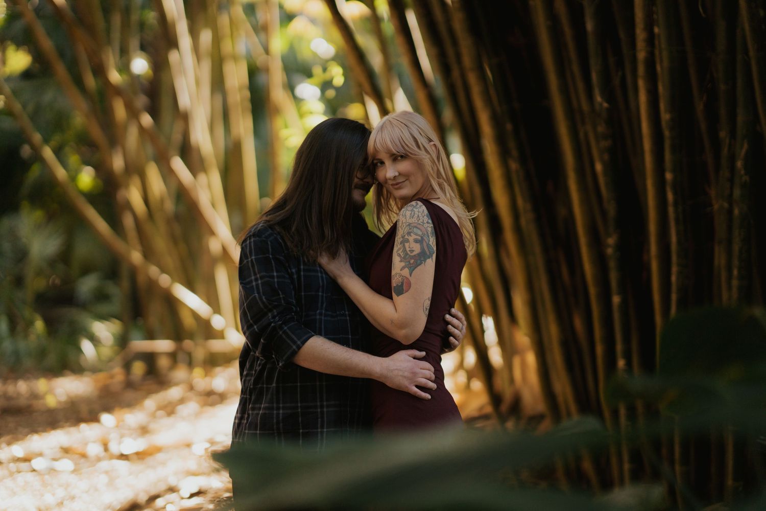 Couple embracing warmly among tall golden bamboo trees in a sunlit tropical garden setting.