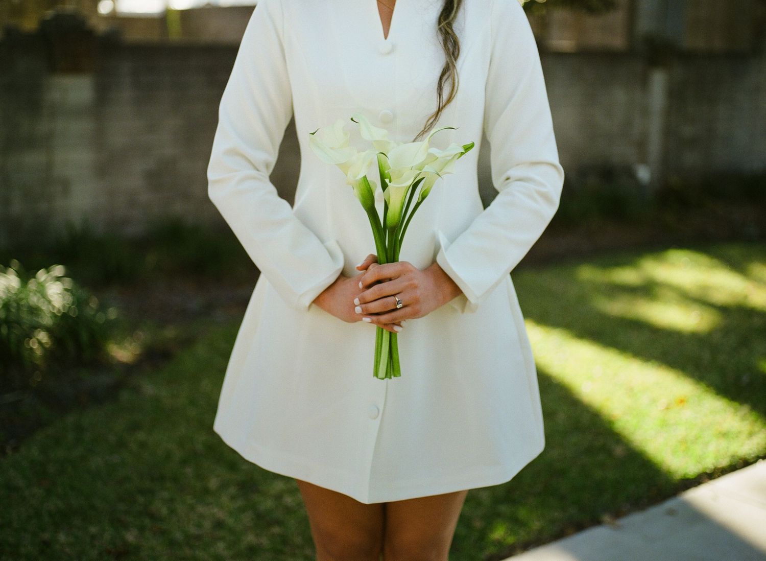 Close-up of bride in white long-sleeve dress holding elegant white calla lily bouquet at her waist.
