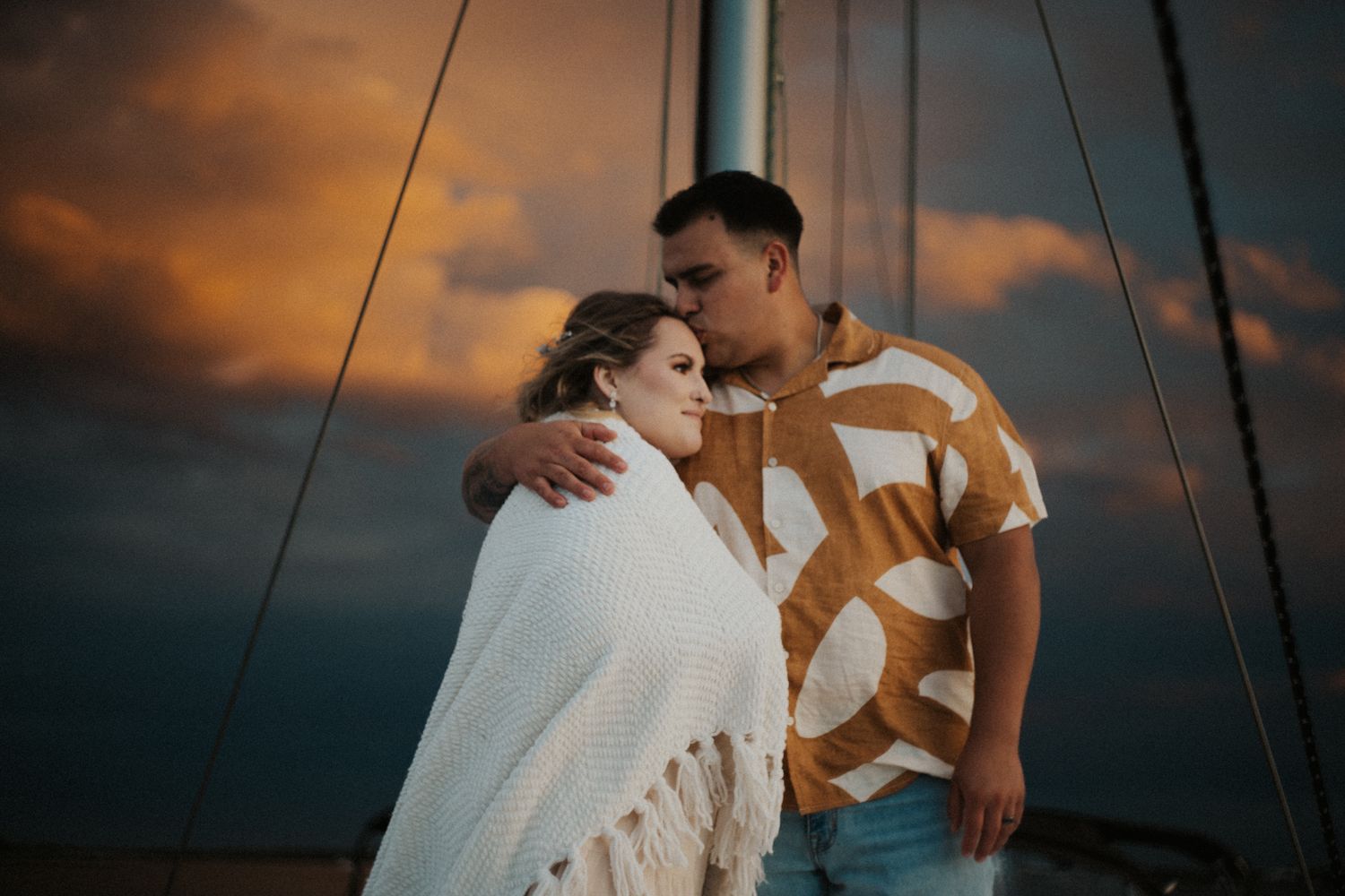 Couple wrapped in white blanket sharing romantic moment on sailboat deck under dramatic sunset sky.