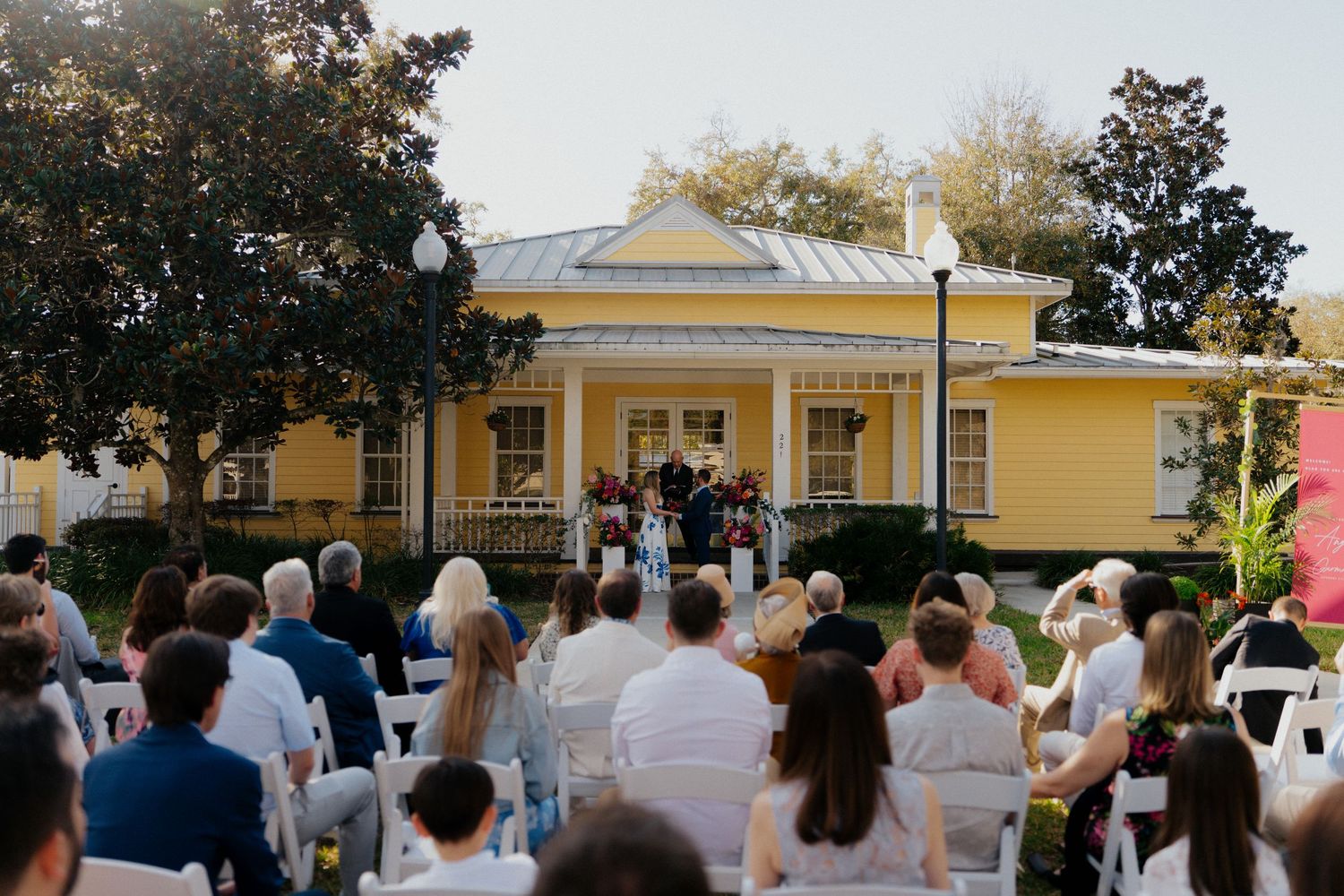 Outdoor wedding ceremony on lawn before yellow historic venue with white chairs and guests in summer attire.