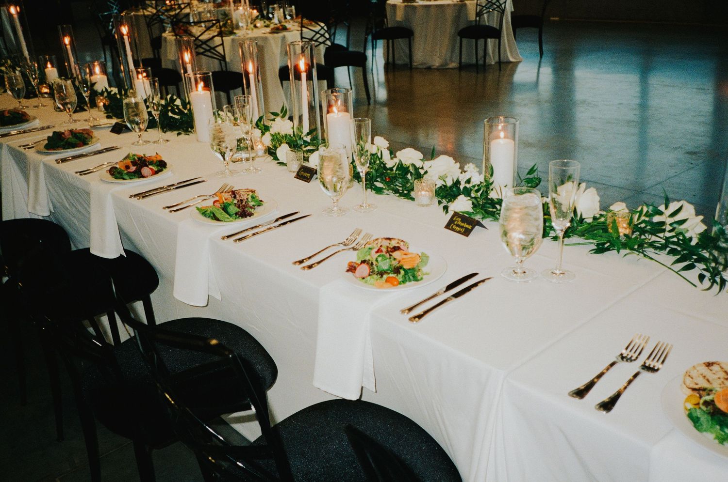 Elegant wedding reception tables decorated with white linens, greenery, and gold accents in a dimly lit venue.