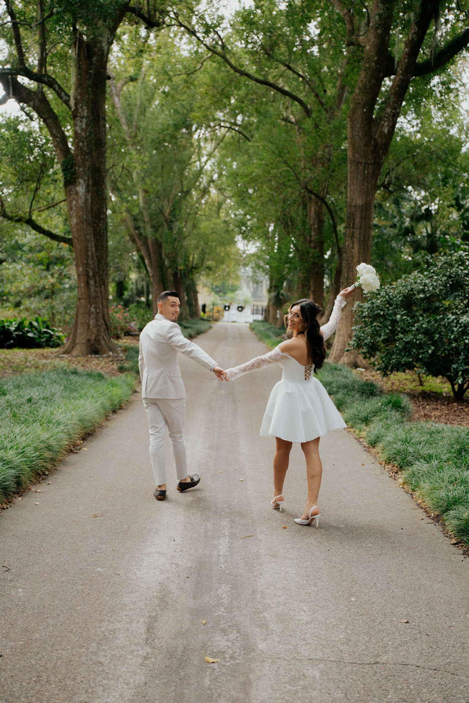 A couple in wedding attire dances playfully along a tree-lined path.