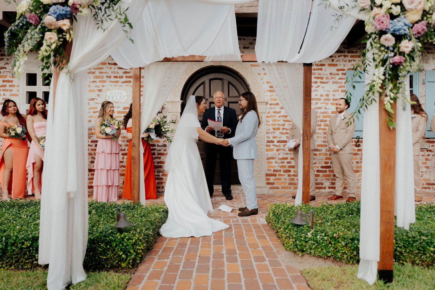 Wedding party poses under brick archways decorated with floral arrangements.