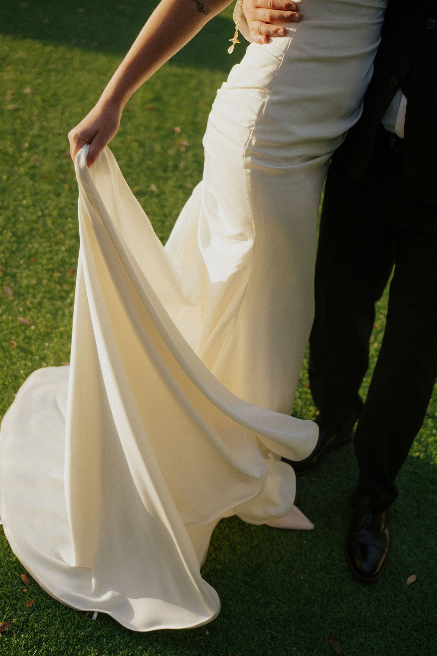Close-up of bride holding flowing white wedding gown train on green grass with groom at Bella Cosa in Lake Wales