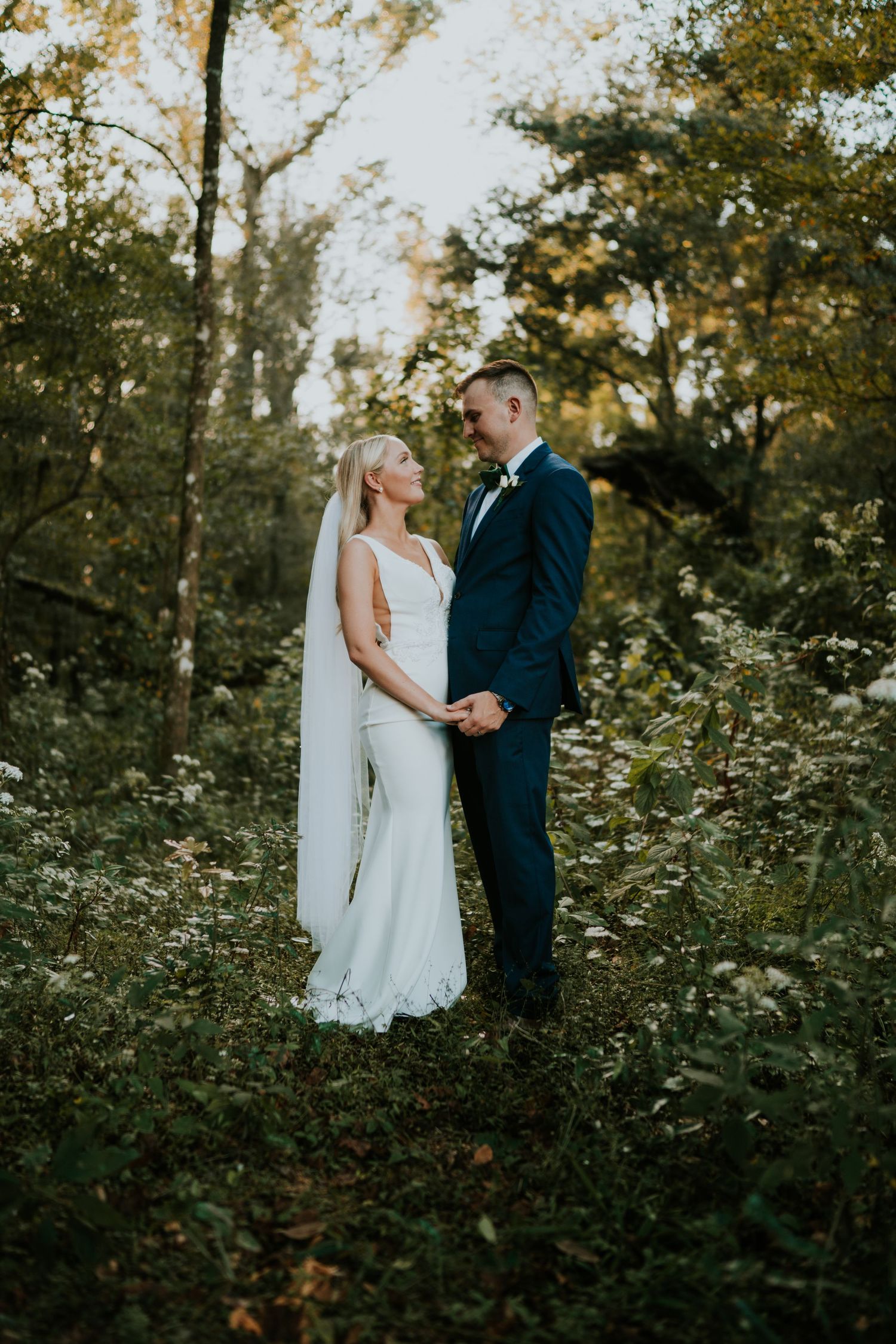 Bride and groom hold hands in a lush forest clearing, she in a white gown with veil, he in a navy suit in new york