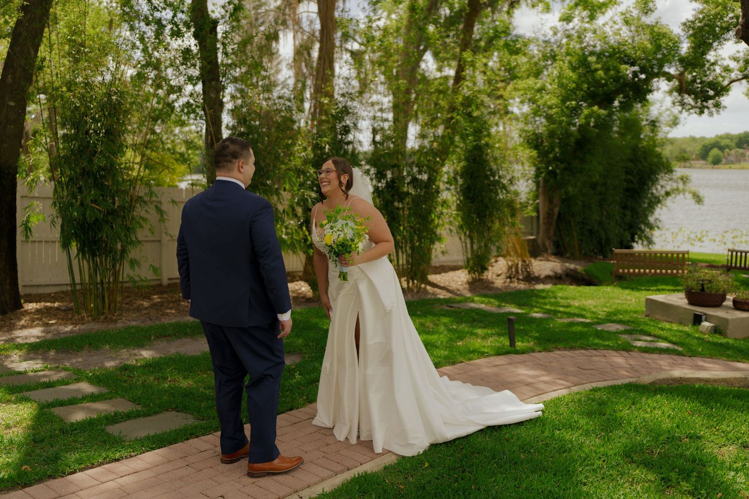 Bride and groom share a romantic first look moment in a lush garden setting near a serene lake.