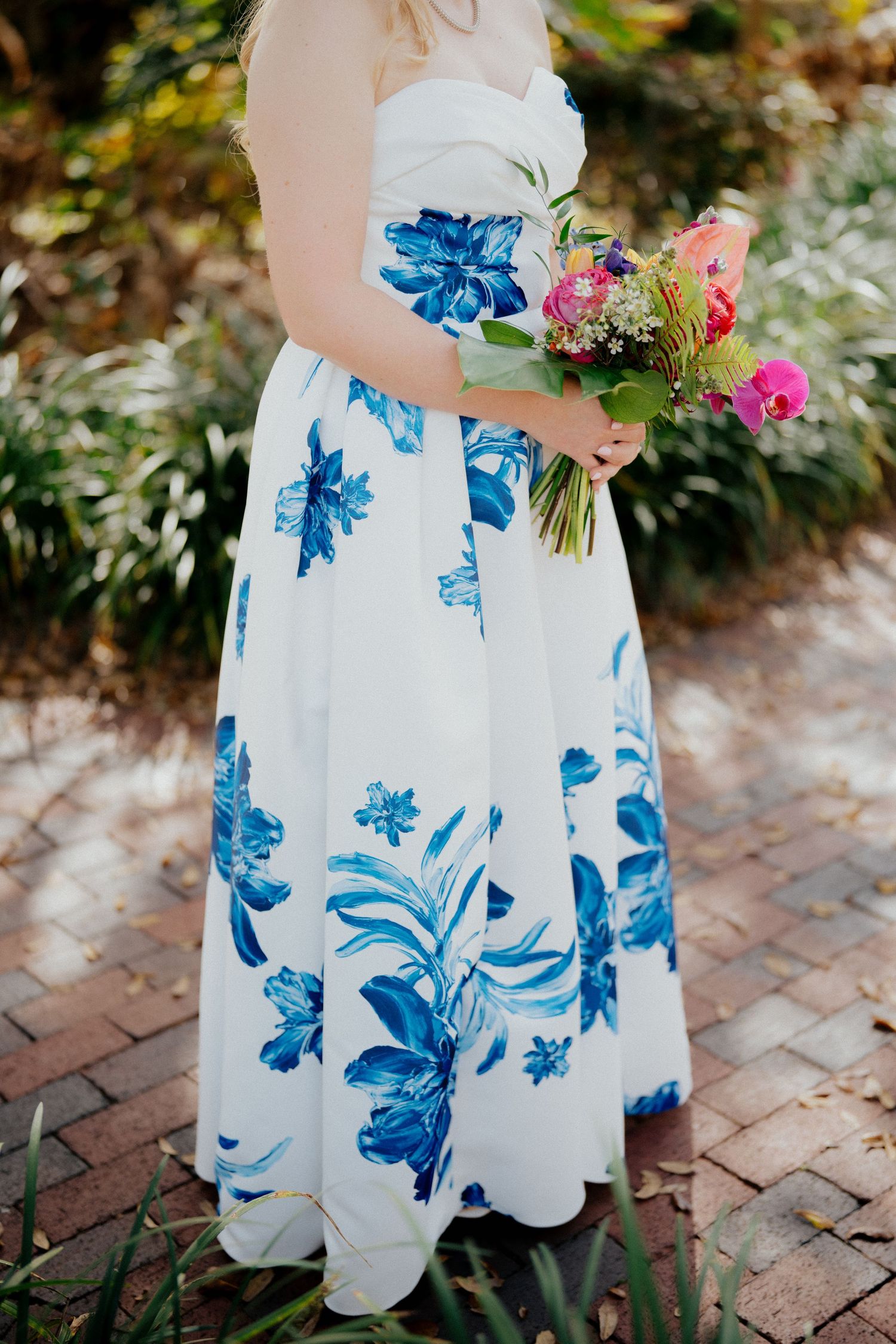 Woman in white and blue floral strapless maxi dress holding colorful bouquet on brick path at micro wedding in Oakland , Florida