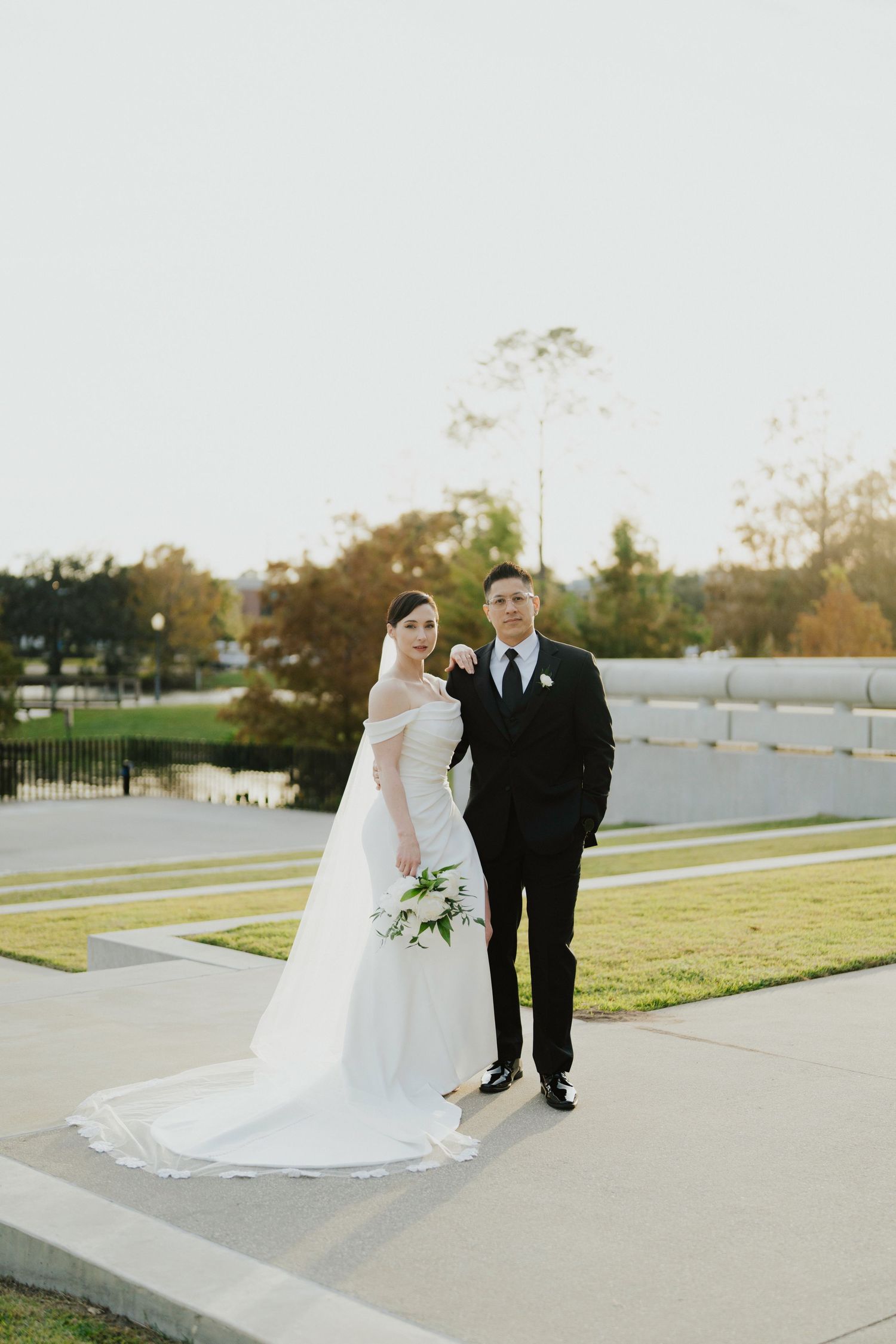 A bride and groom pose together on a paved walkway during their outdoor wedding photoshoot at sunset.