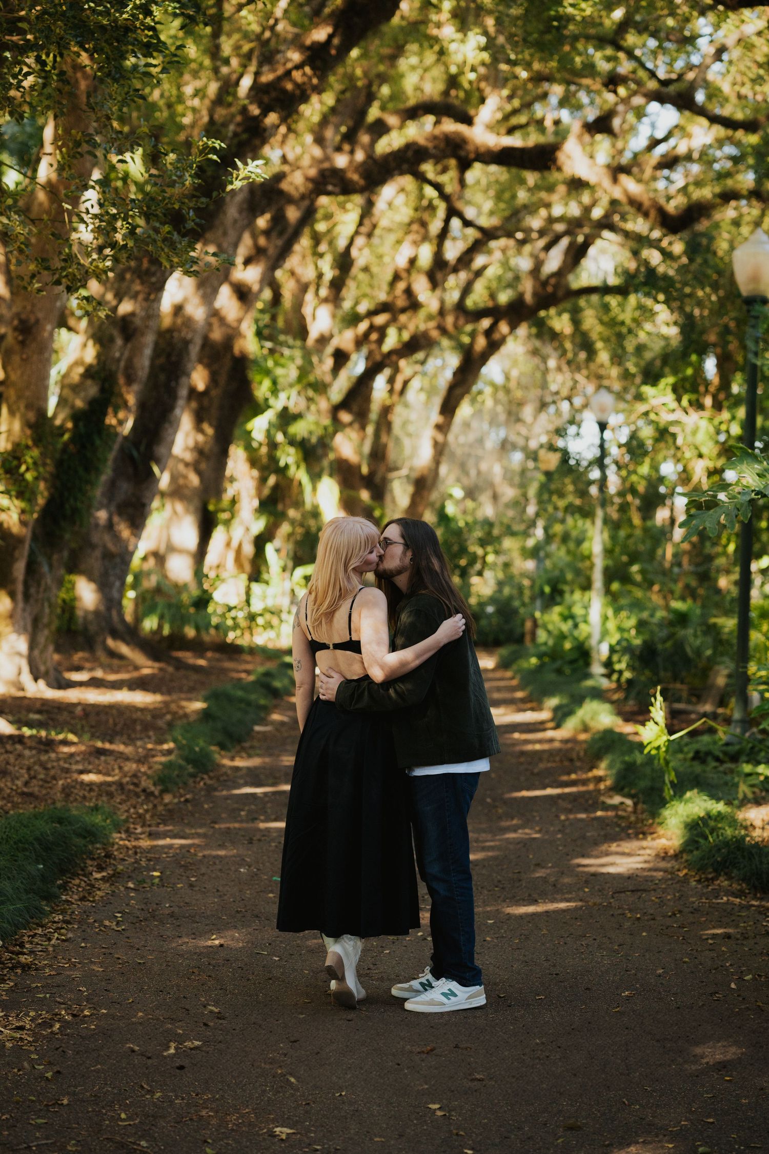 A romantic couple shares intimate moments along a tree-lined dirt path in a sunlit forest setting.
