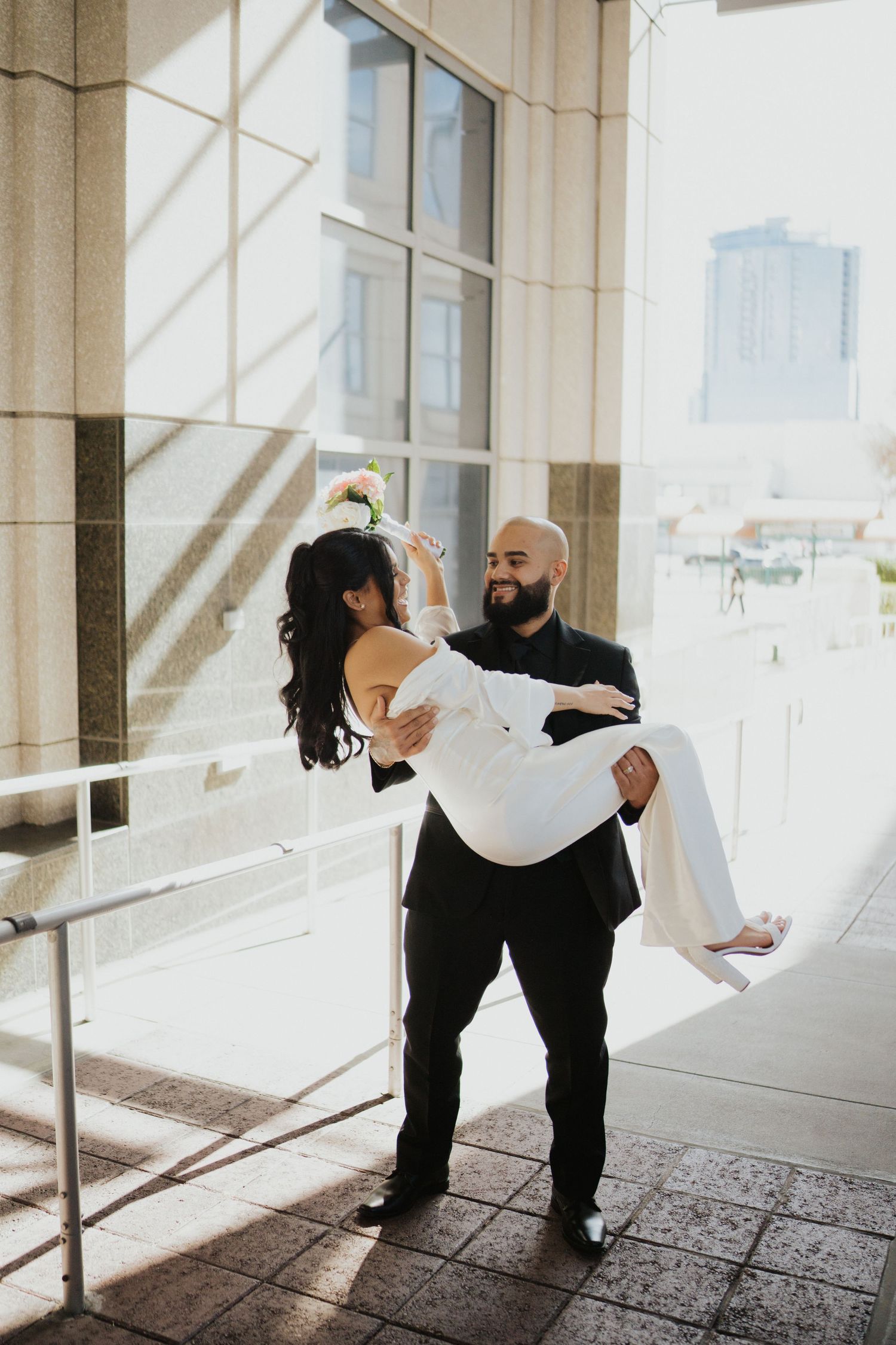 A romantic moment captured as a groom carries his bride in a white dress near a building entrance.