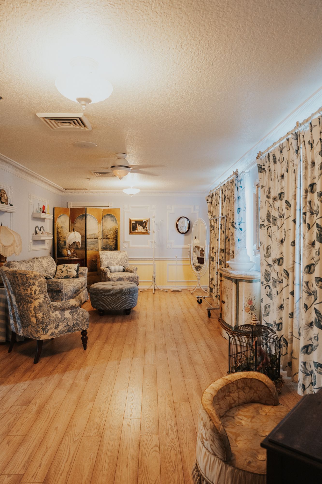 Long view of an elegant sitting room with vintage furniture, floral curtains and hardwood floors.