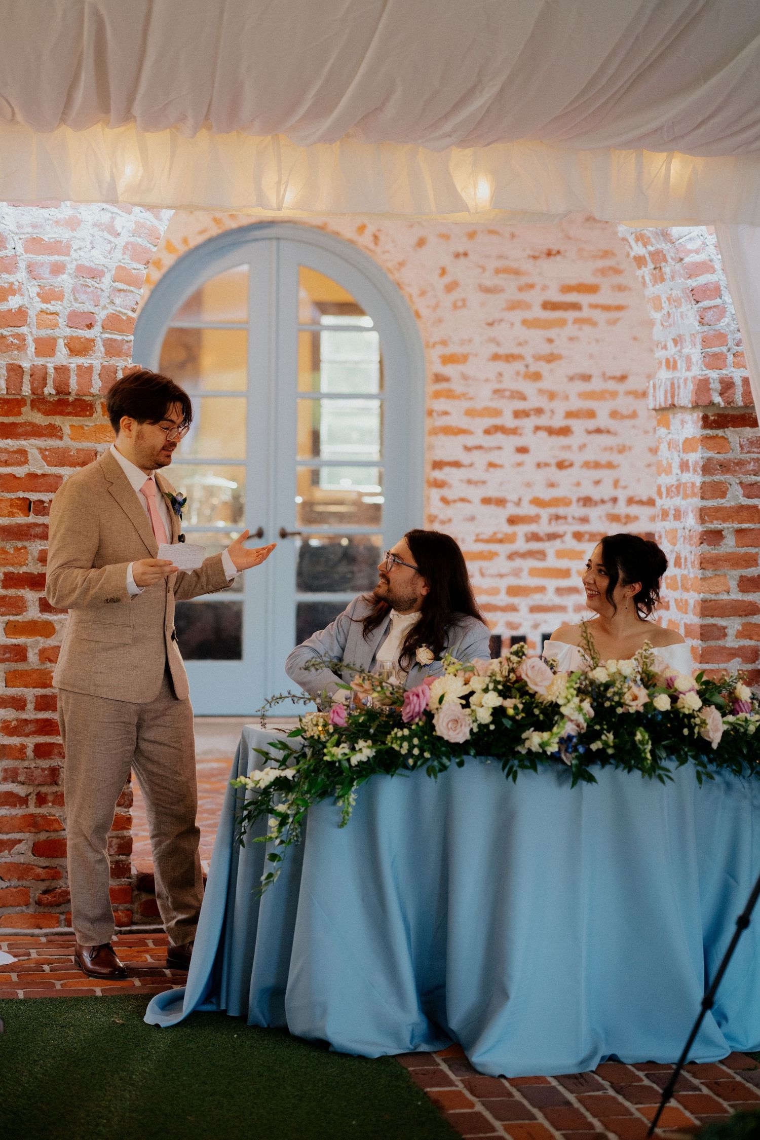 Wedding reception head table decorated with light blue linens and floral arrangements against exposed brick walls.