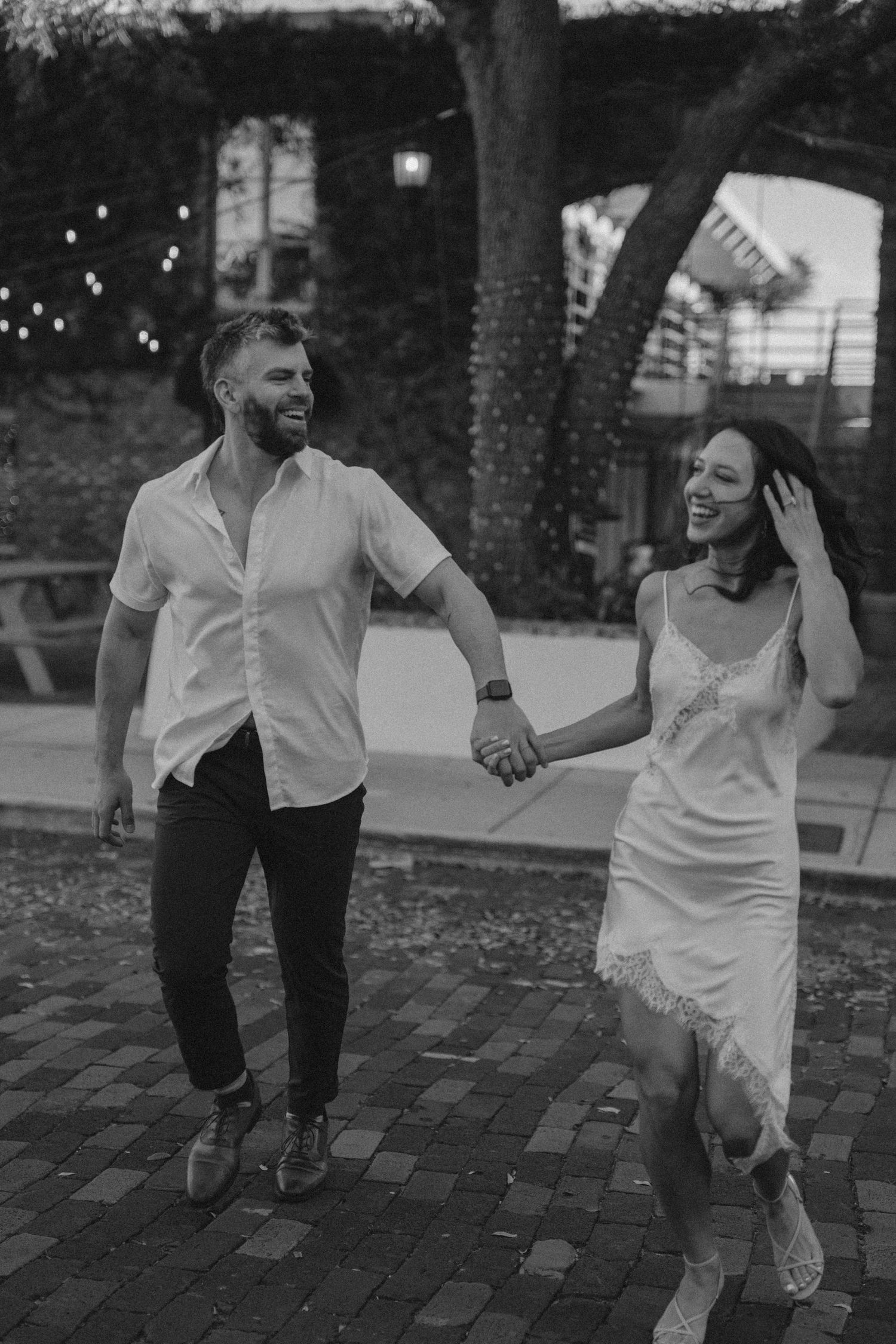 Black and white photo of laughing couple holding hands running on brick path with string lights above.