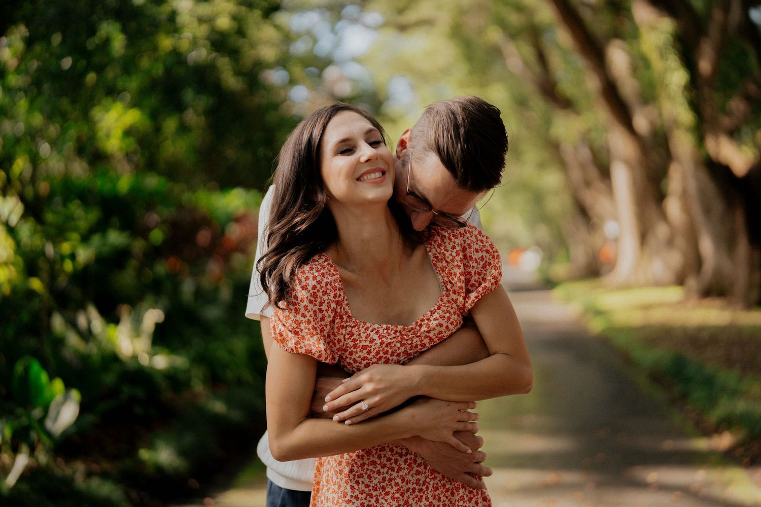 Man hugs woman from behind on a sunny tree-lined path, woman smiling in orange floral dress.