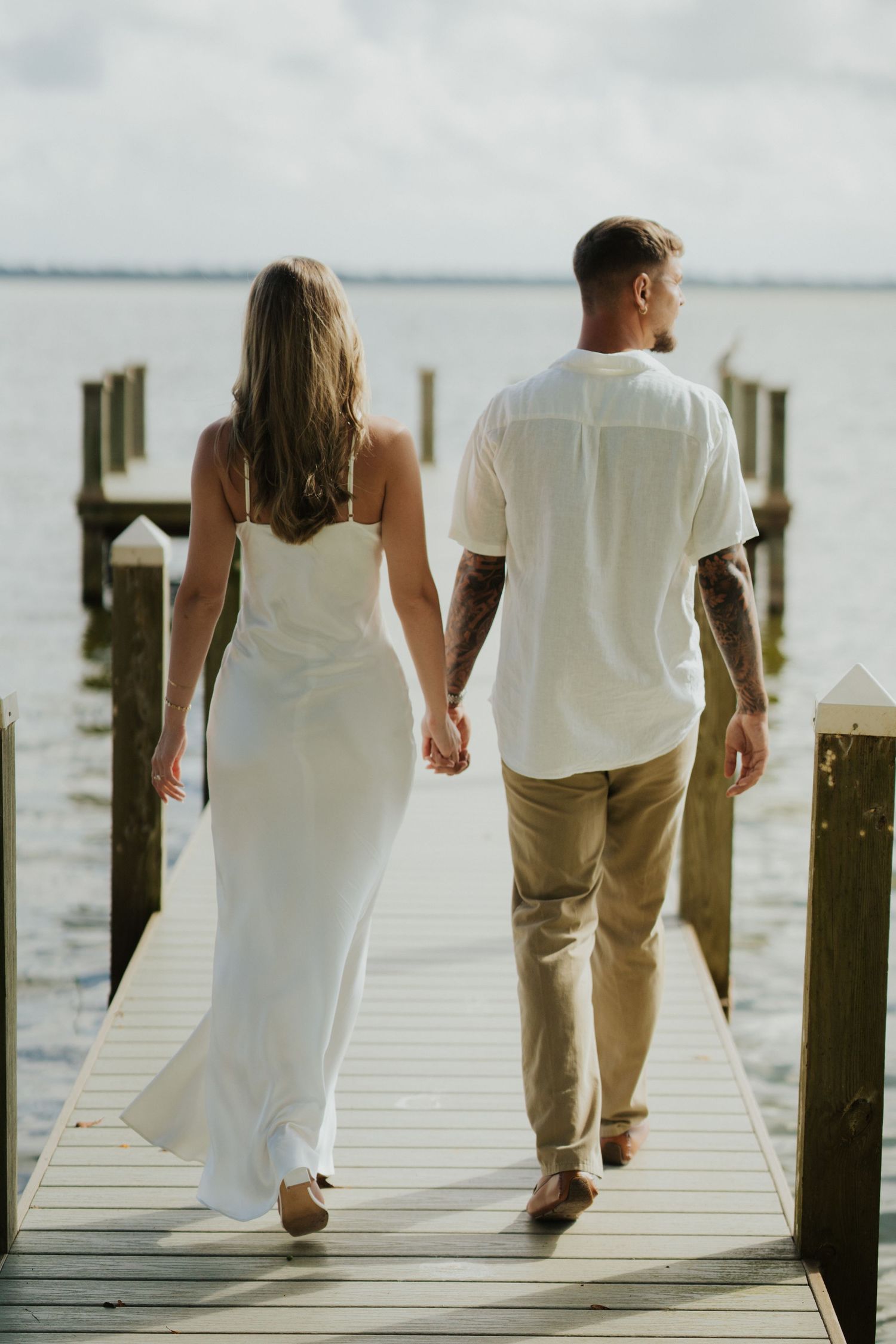 A couple in white and khaki walks hand in hand along a wooden dock by the waterfront.