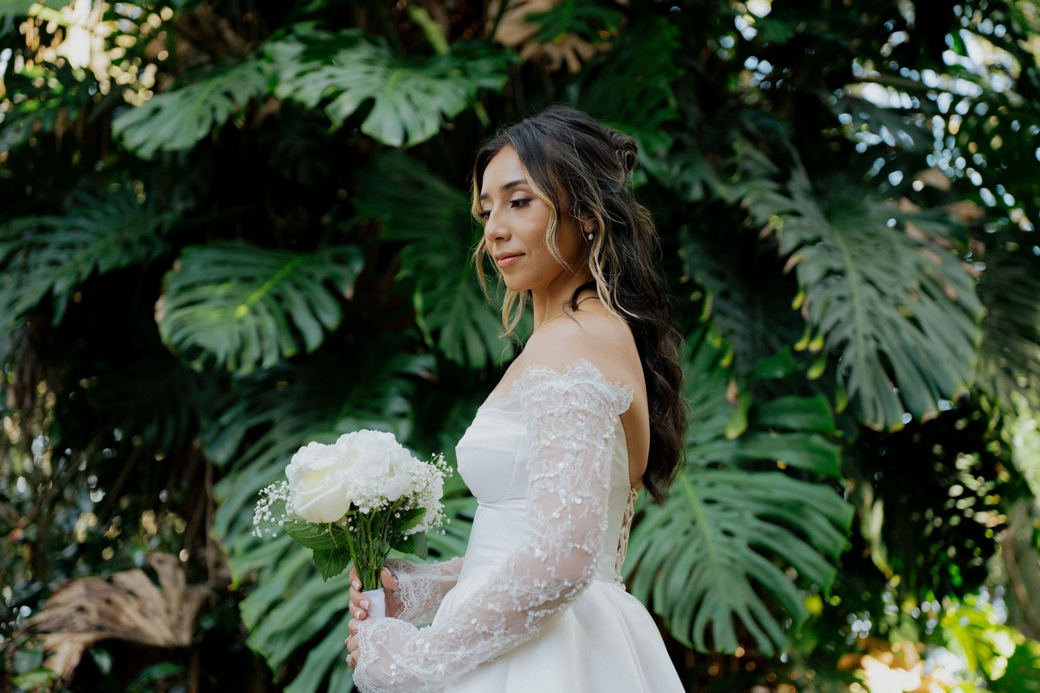 Bride in lace wedding dress poses elegantly in a lush tropical garden setting with monstera leaves.