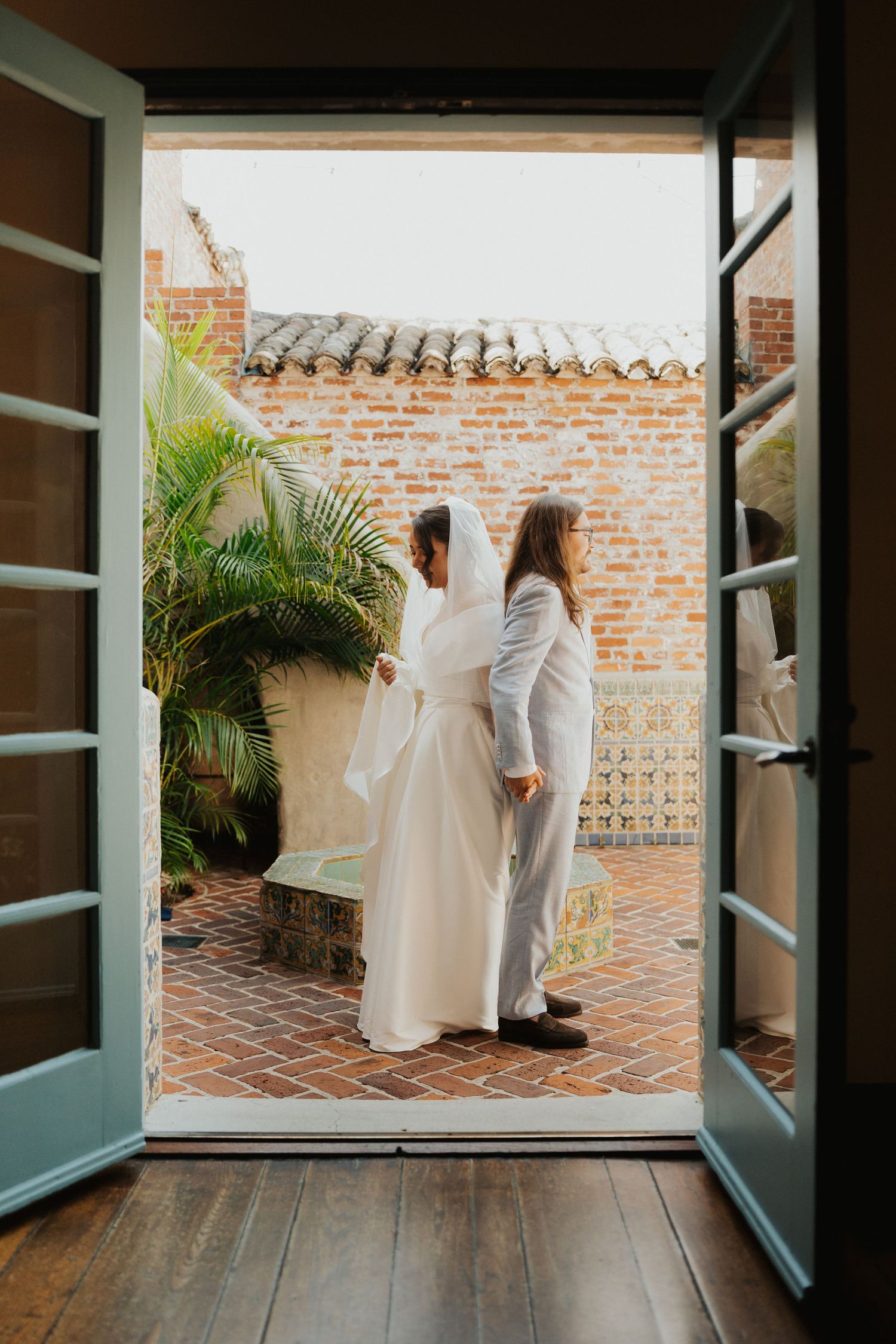 Wedding party walking through French doors onto a tiled courtyard with brick walls and tropical plants.