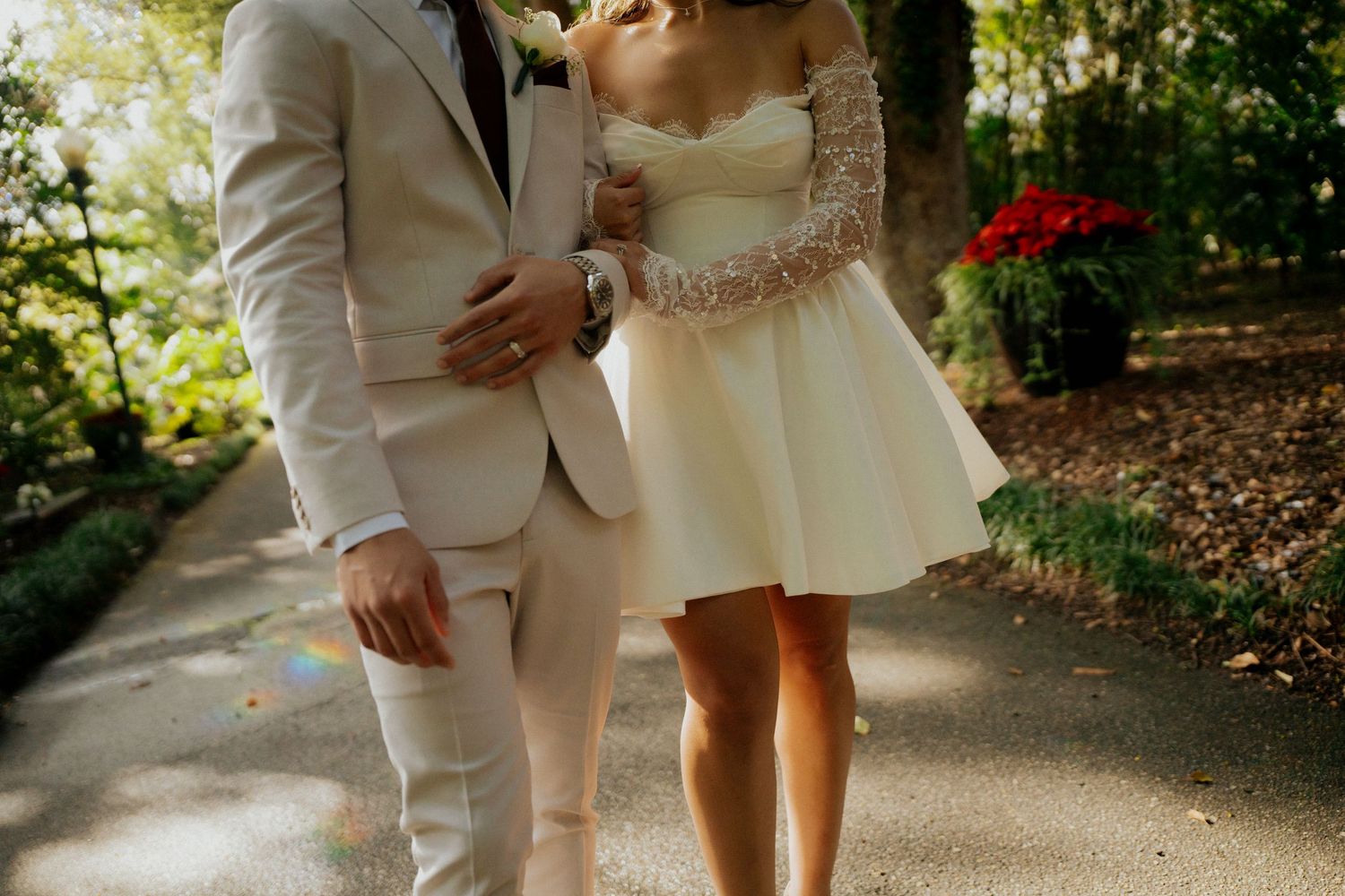 Couple in light-colored wedding attire walking together on a sunlit garden path.