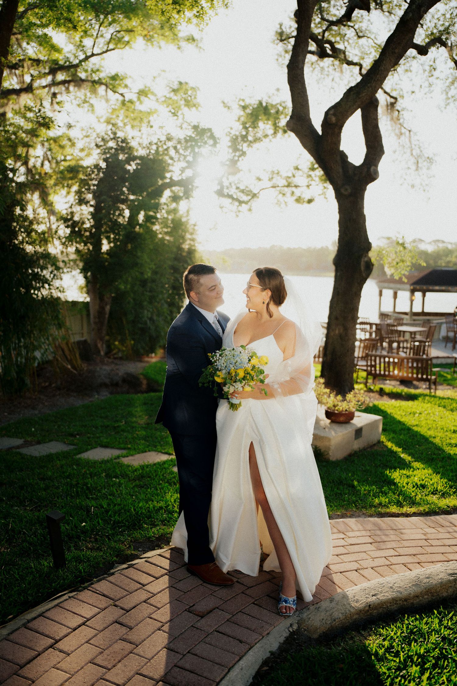 Bride and groom share a romantic moment on a brick path by a sunlit lakeside garden, bride holding yellow and white bouquet.