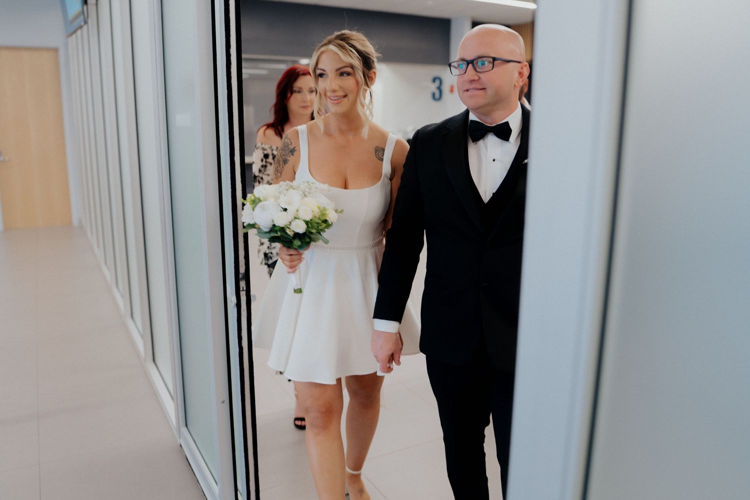 Bride in short white dress holding bouquet walks with groom in tuxedo down a bright hallway.