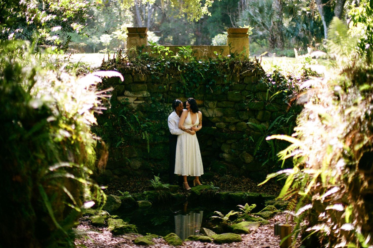 Couple in white attire embracing on a stone bridge surrounded by lush tropical greenery.
