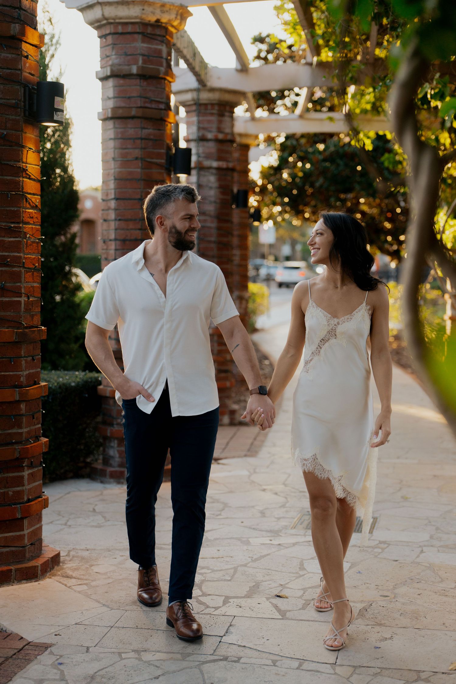 Stylish couple holding hands and walking along a sunlit brick colonnade pathway at golden hour.