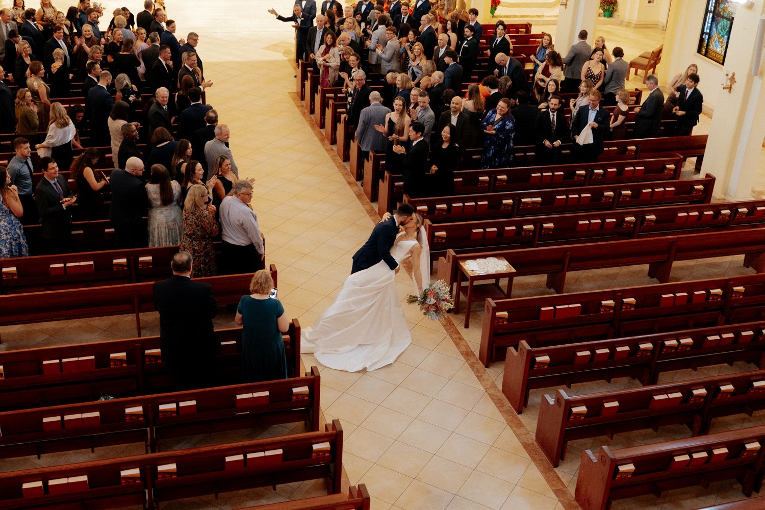 A wedding ceremony procession walking down the aisle of a traditional church with wooden pews and gathered guests.