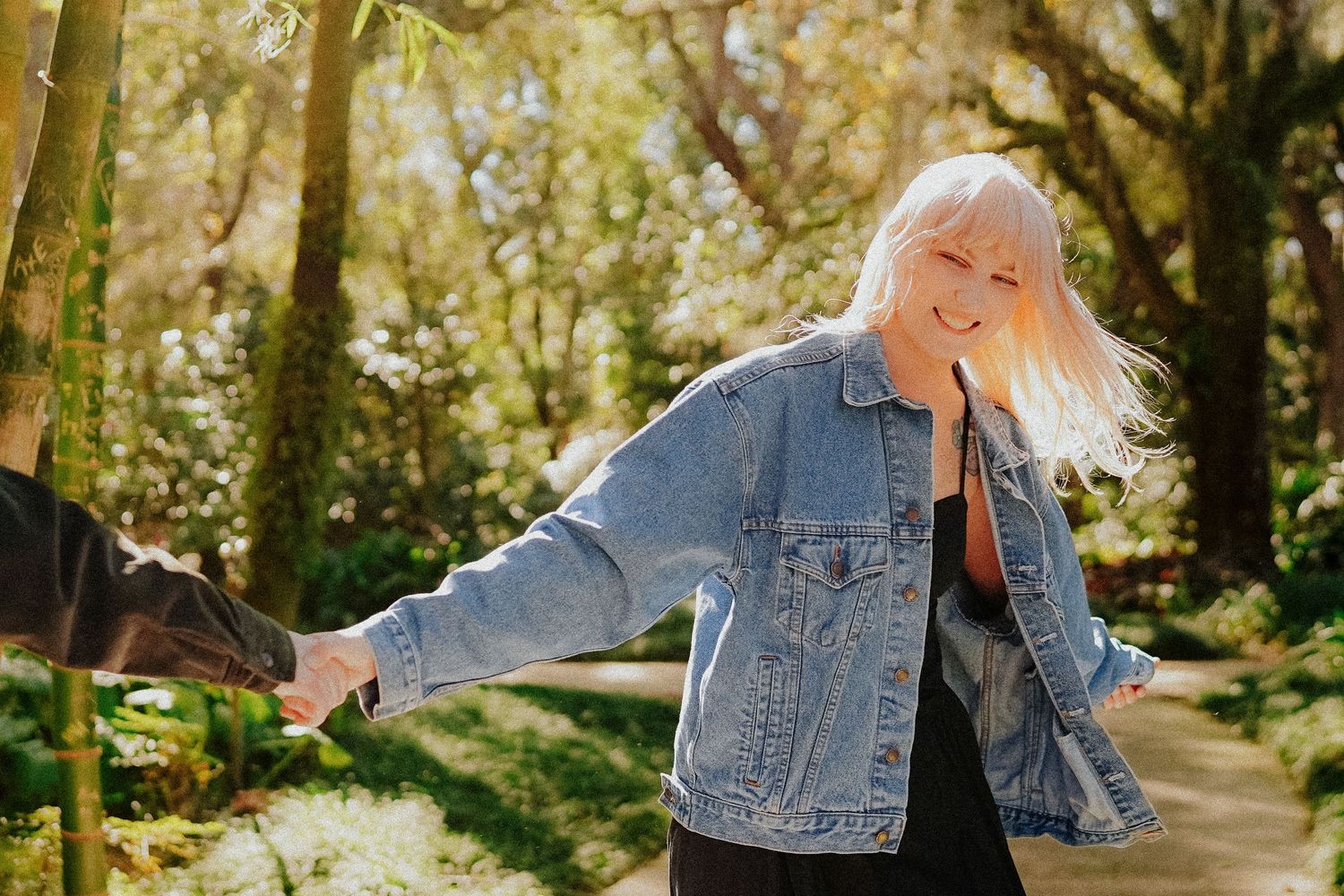 Someone with light hair dances joyfully on a sunlit forest path wearing a denim jacket.