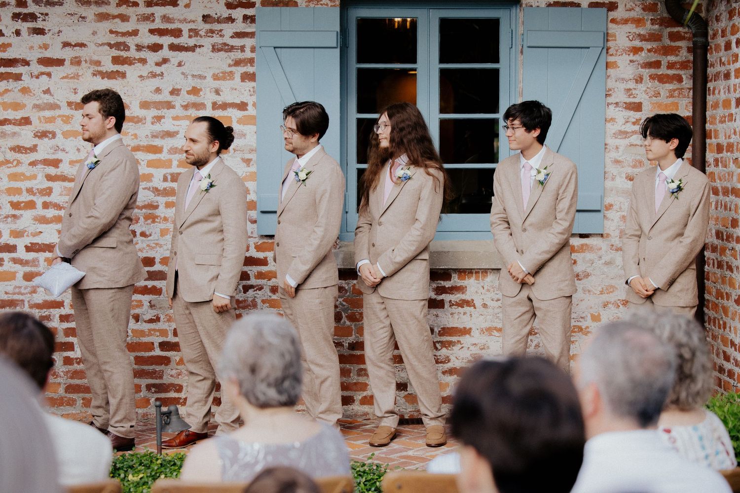 Wedding party poses in front of a brick building with light blue shutters in coordinated formal attire.