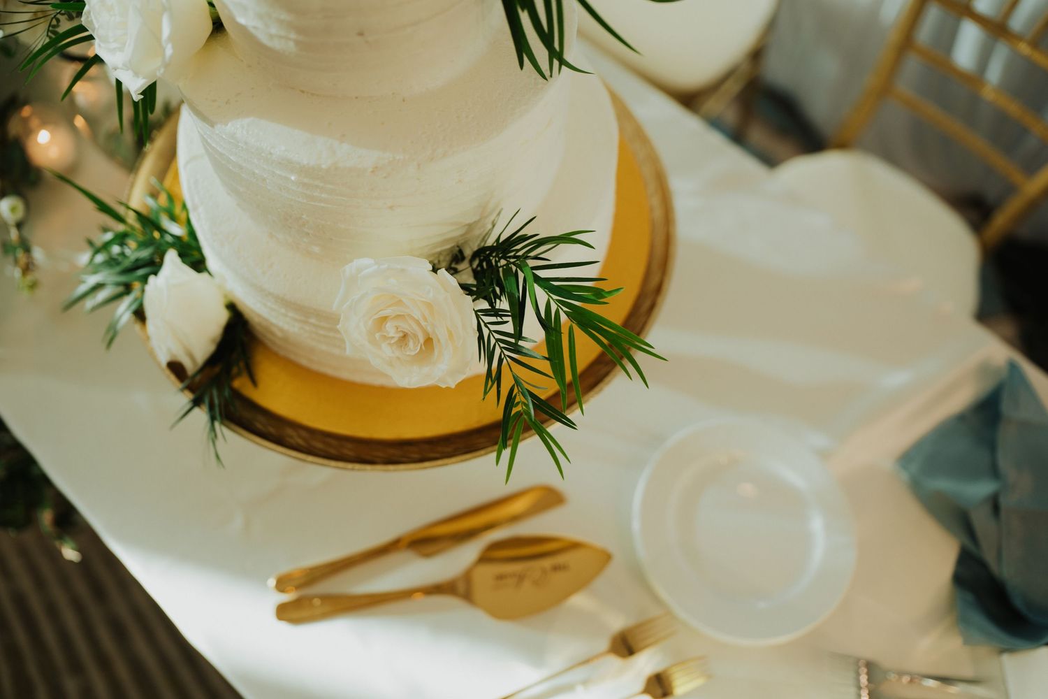 Three-tier white wedding cake decorated with roses and greenery on a gold stand with wooden cake servers.
