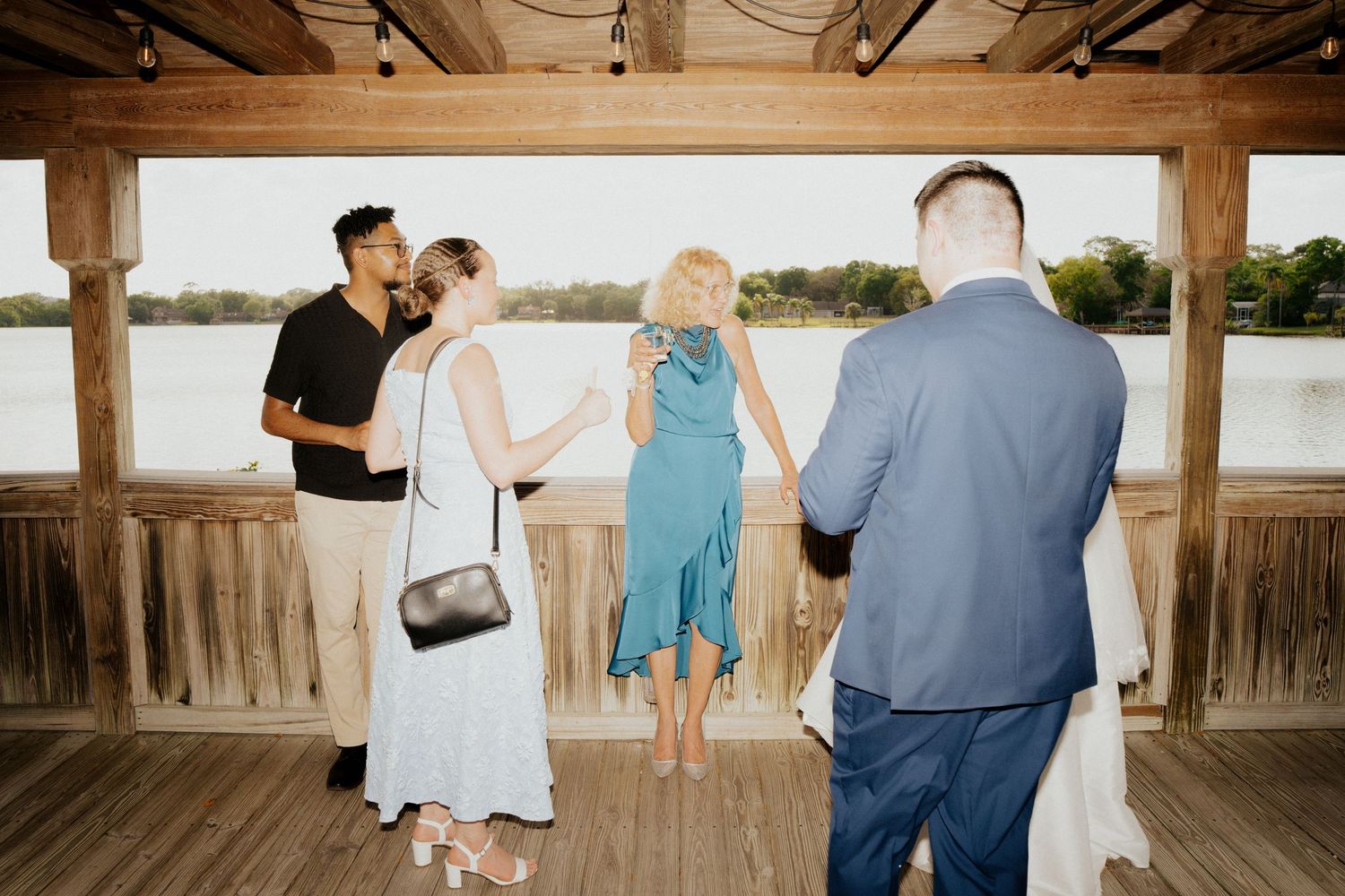 Wedding guests socializing on a rustic wooden waterfront pavilion at sunset reception.